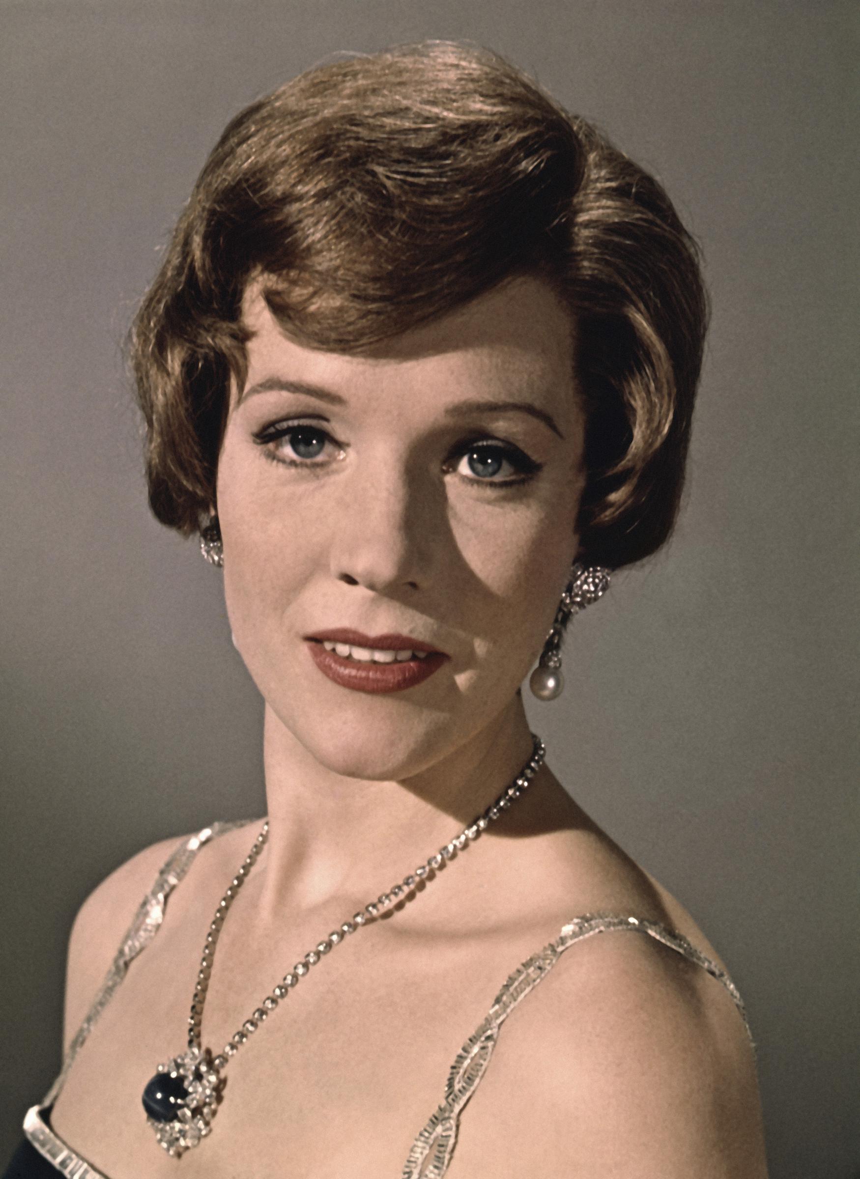 Julie Andrews exudes classic Hollywood glamour in a promotional portrait for the 1970 musical drama "Darling Lili," directed by Blake Edwards. The English actress and singer wears a sophisticated sequin-trimmed gown paired with a diamond and sapphire pendant necklace, pearl drop earrings, and elegant winged eyeliner framing her waved brunette coiffure.