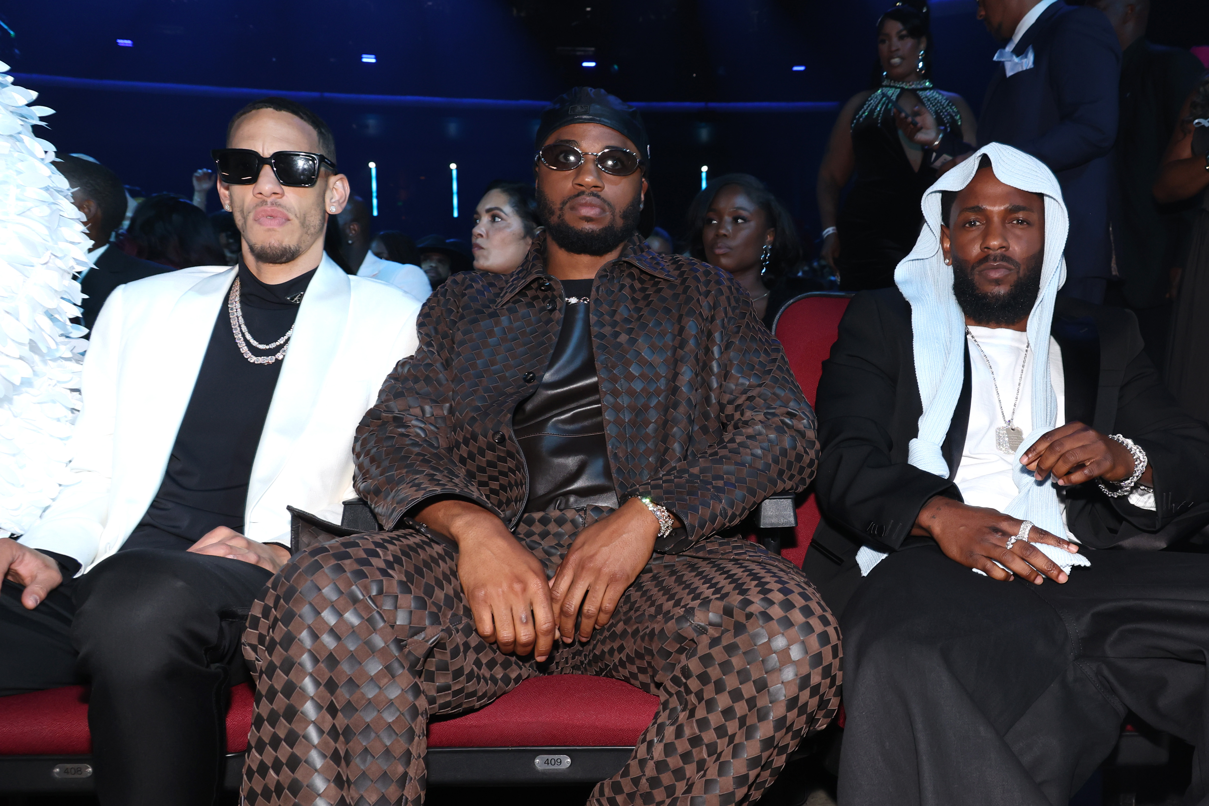 Seated front row at the 2025 BET Awards, Kendrick Lamar poses with fellow attendees, King Matta and Dave Free, in a composed, relaxed moment. He sported his now-viral headpiece alongside a sleek black suit and white shirt.
