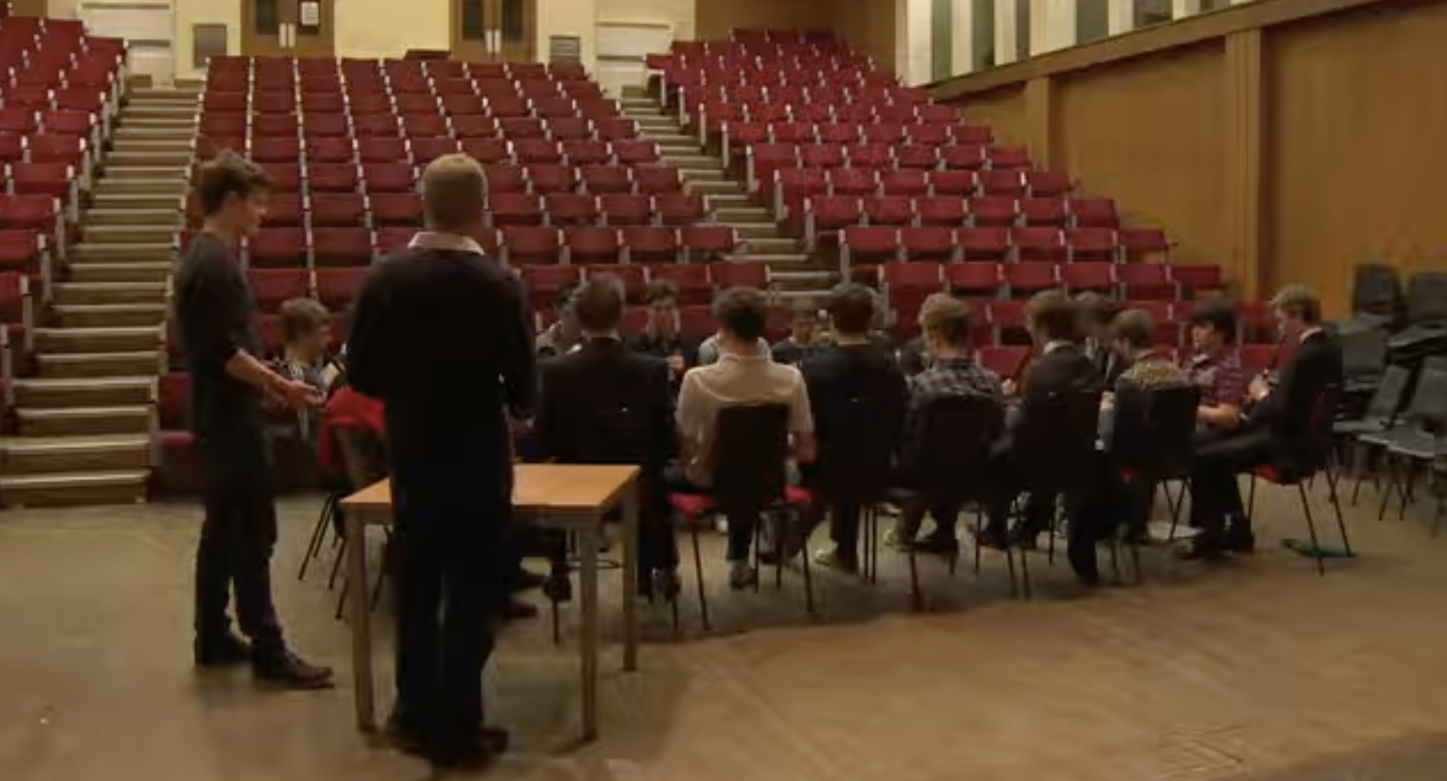 Students sit in a group on the floor of a large hall surrounded by rows of red tiered seating. The gathering offers a glimpse of the shared spaces used for activities at Eton College. | Source: Youtube/Paradoxyy
