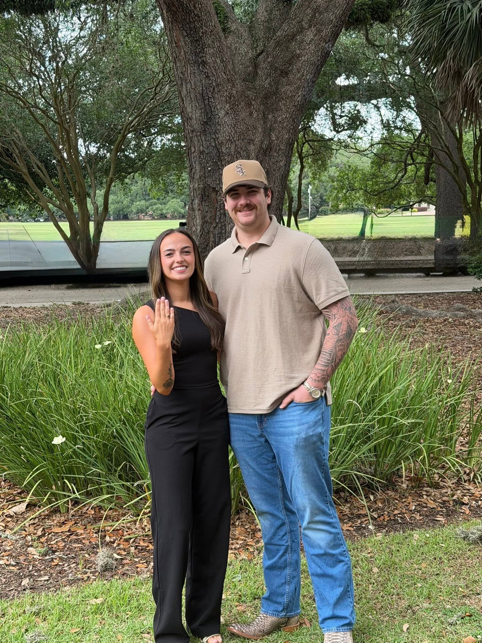Shane David Hirstius and Leah Victoria Fonte pose outdoors by a large oak tree to celebrate their engagement. Leah proudly shows off her new engagement ring while smiling in an elegant black sleeveless jumpsuit, standing beside Shane, who smiles in a tan polo shirt, blue jeans, cowboy boots, and a baseball cap. | Source: Facebook/StevenMarjorie Hirstius