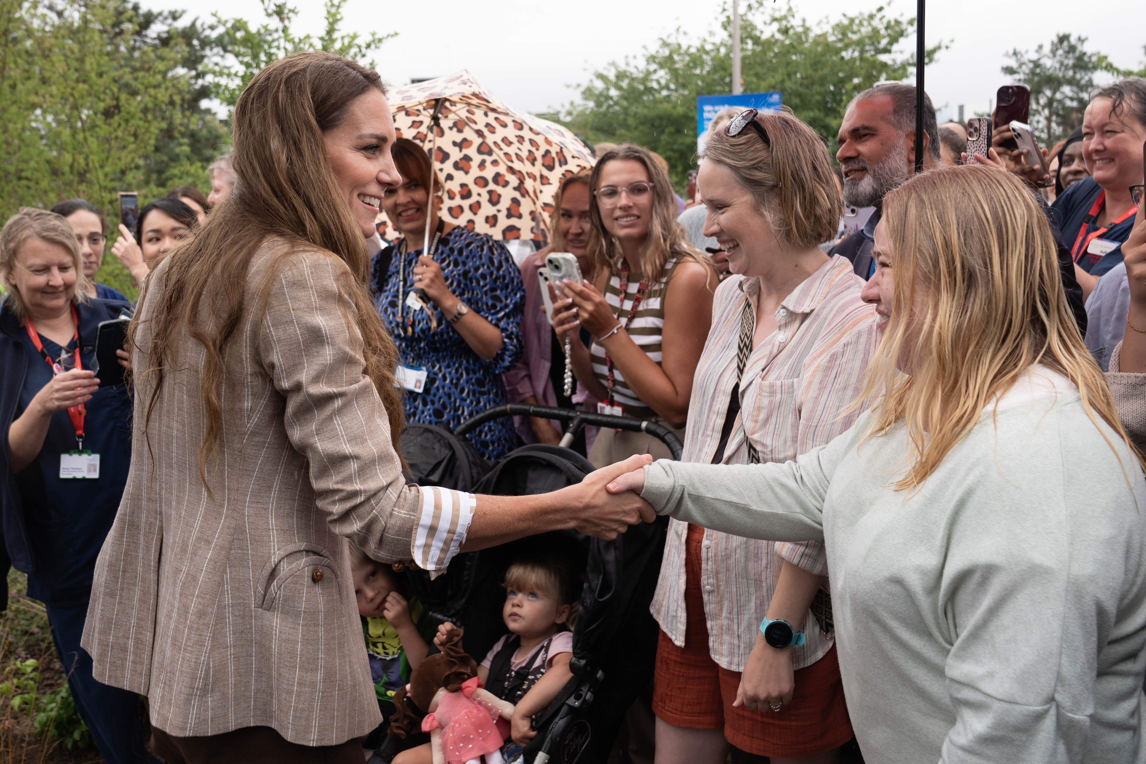 In a candid exchange, Princess Catherine greets well-wishers with a handshake and a smile. The crowd, including children in strollers, eagerly gathered to meet the royal during her public engagement.