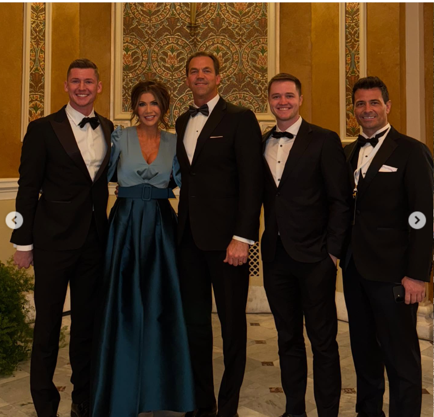 Kristi and Bryon Noem pose with a group of men in formal attire, all smiling as they stand together inside an ornate, warmly lit interior. | Source: Instagram/kristinoem