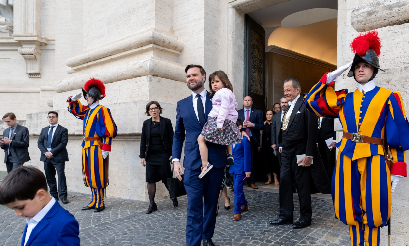 JD Vance walks out of a stone building while carrying one of his young children, flanked by Swiss Guards in traditional uniforms who stand at attention on either side of the entrance. | Source: Instagram/vp