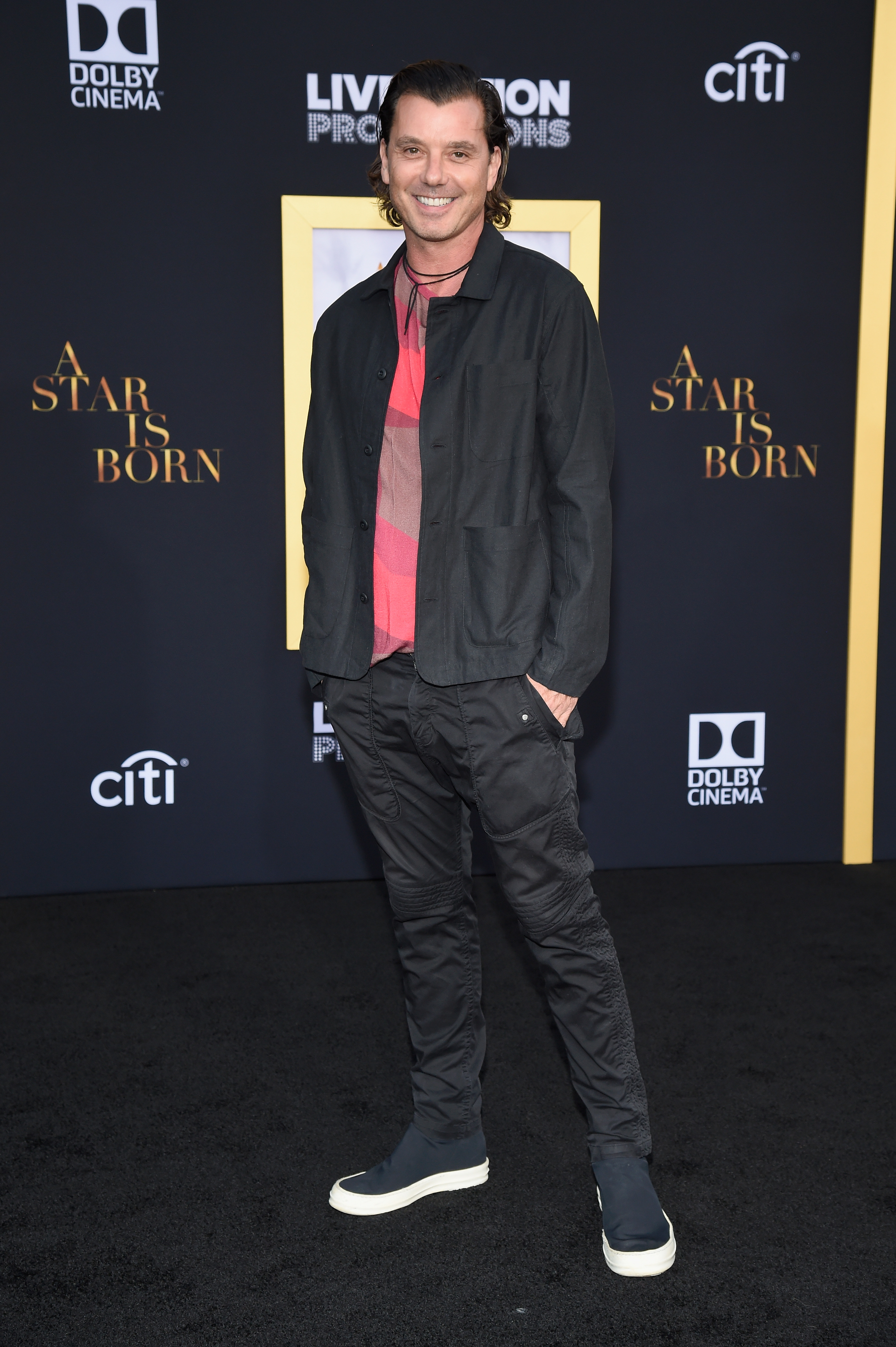 Gavin Rossdale smiles at the Los Angeles premiere of "A Star Is Born," wearing a black jacket, pink shirt, dark pants, and slip-on sneakers.