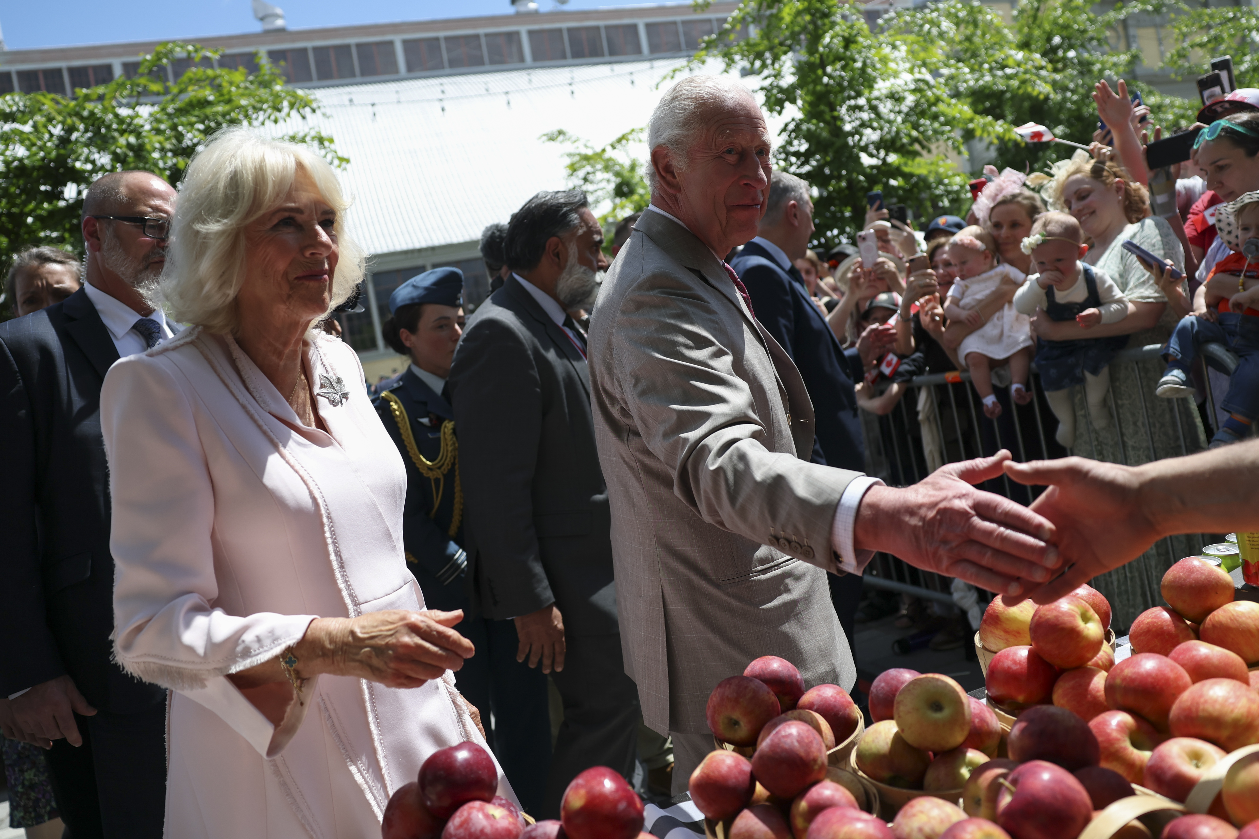 King Charles III and Queen Camilla greet a vendor at Lansdowne Park, stopping by a stall with apples neatly displayed, adding a friendly touch to their community visit.