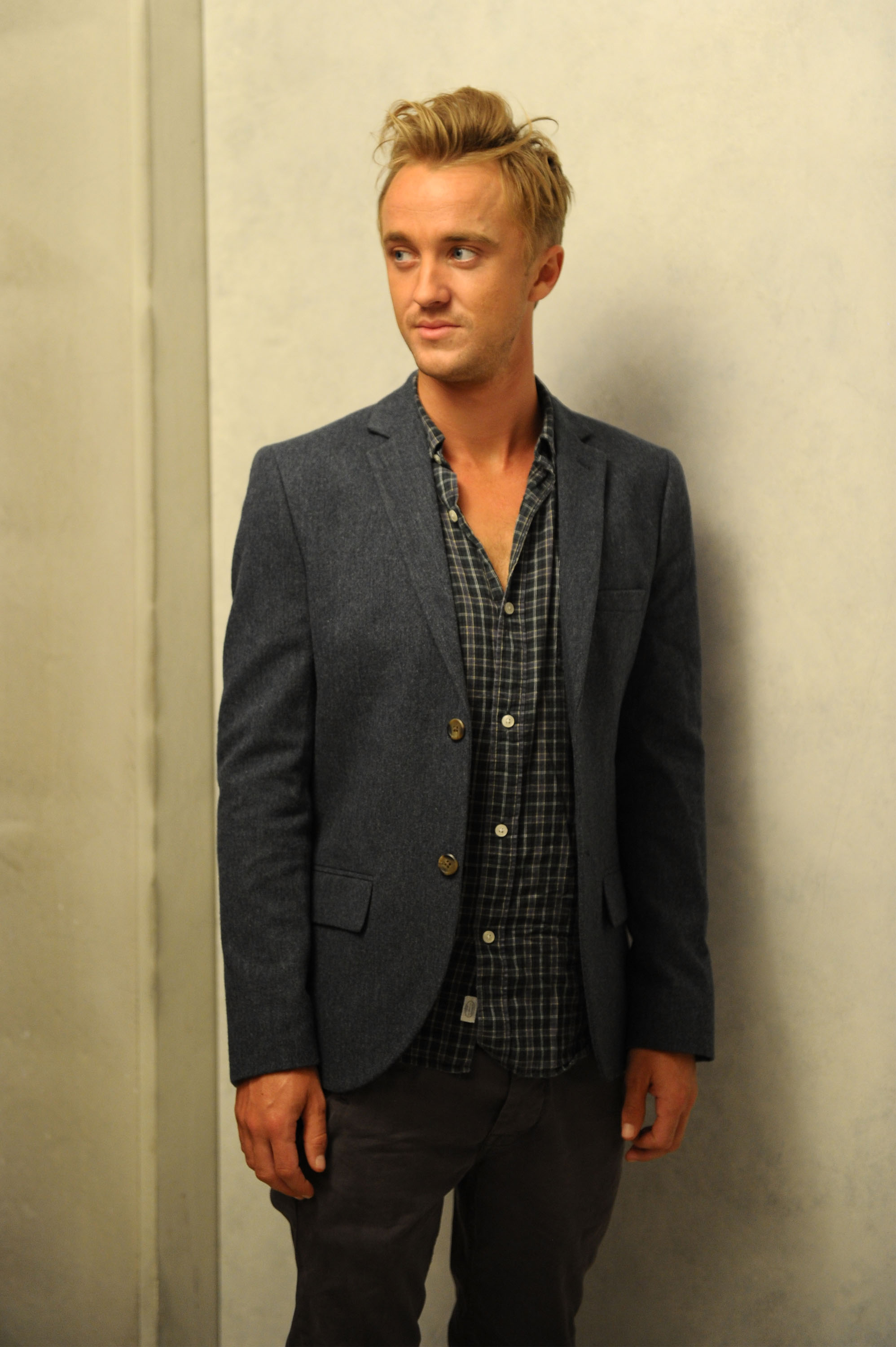 Tom Felton stands in a softly lit studio, blending casual ease with tailored polish in a checked shirt and blue blazer. His relaxed pose and thoughtful glance lend a quiet charm to the Toronto International Film Festival's bustling atmosphere. Amid the event's cinematic excitement, he brings calm, understated confidence.