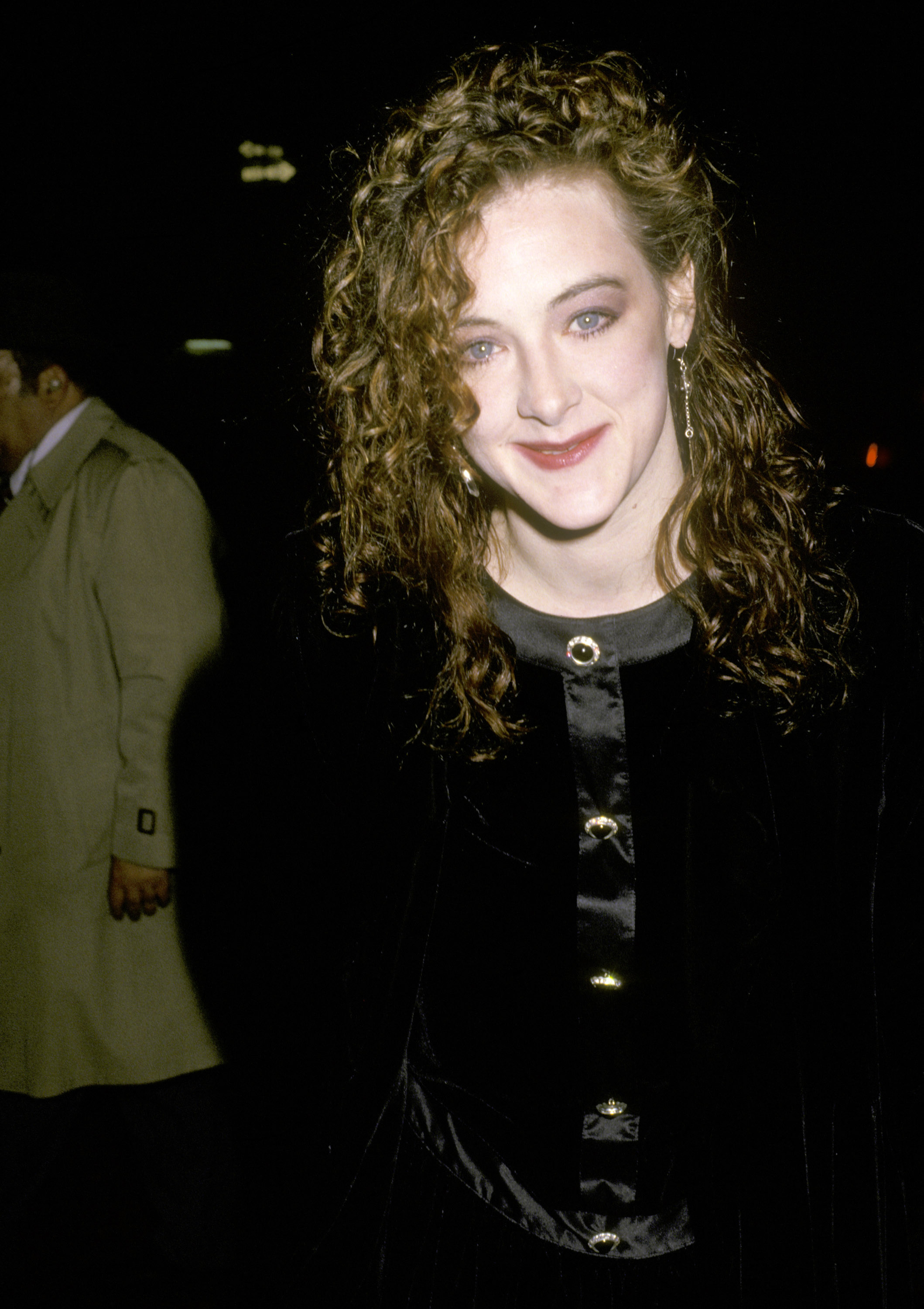 Wearing soft curls and a black ensemble with satin details, Joan Cusack smiles warmly for the camera. Her understated look highlights her youthful features and bright eyes.