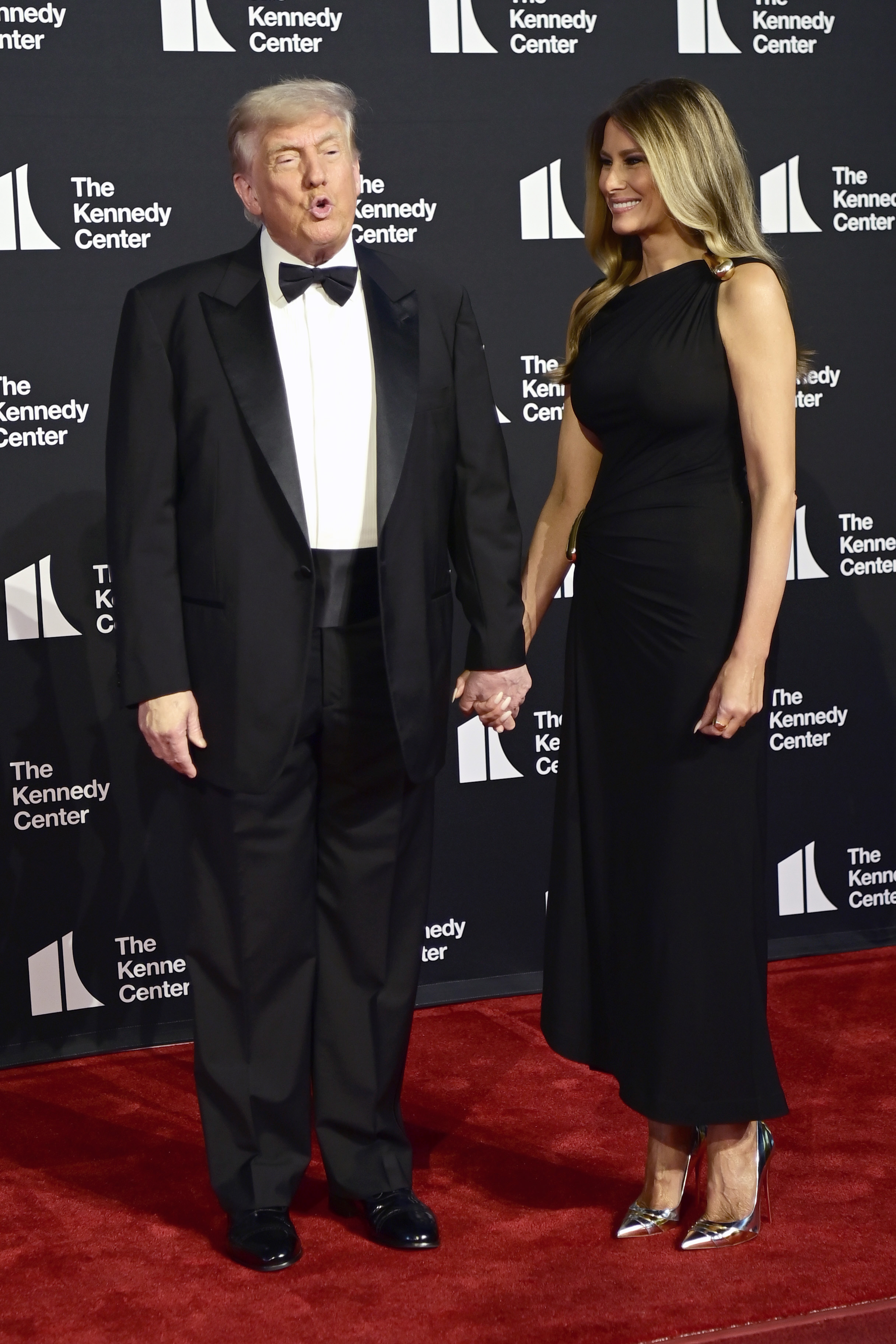 In a playful mid-expression, the president holds hands with the first lady as she looks on with a smile, capturing a spontaneous moment on the red carpet at the Kennedy Center.