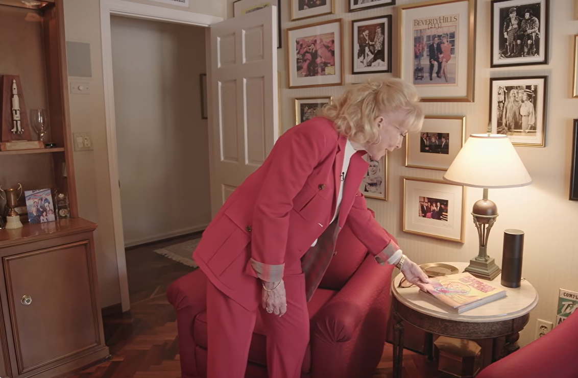 Barbara Eden bends over a small round side table beside a red armchair to examine a colorful book, with a lit table lamp and a wall of framed photographs adding warmth to the cozy sitting area. | Source: YouTube/People