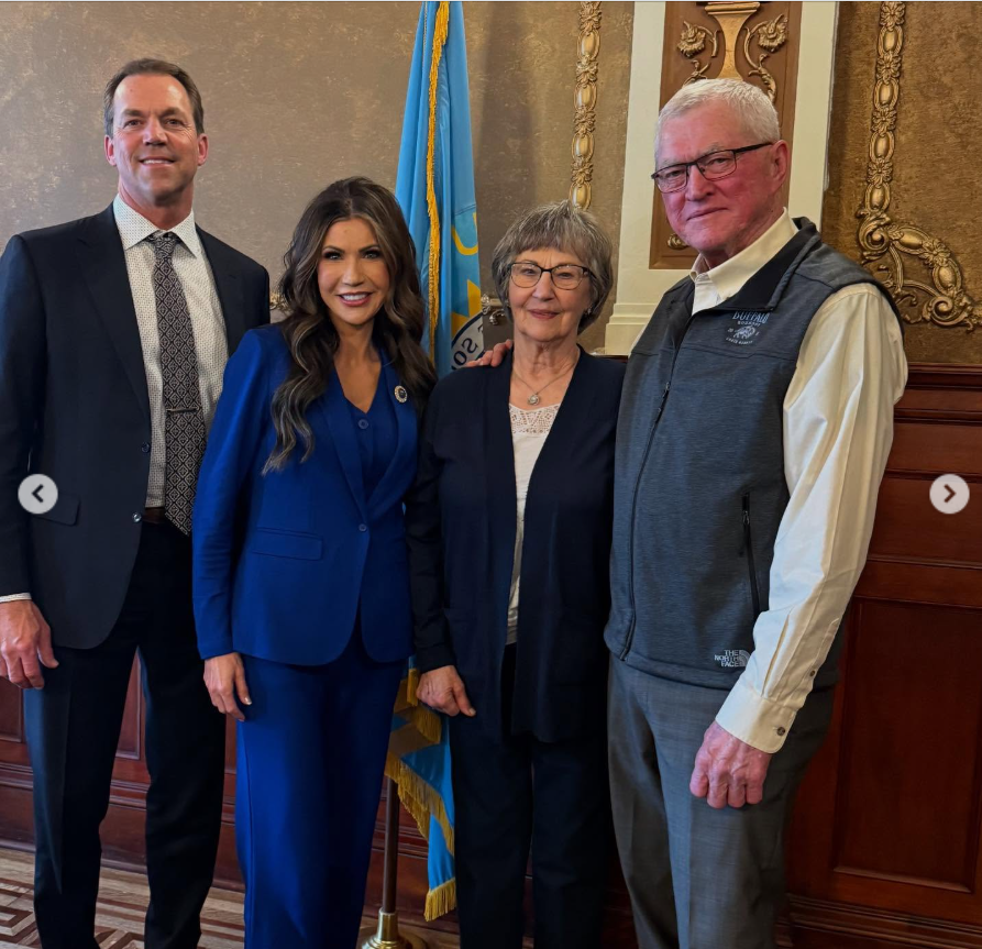 Bryon and Kristi Noem stand alongside two older adults in a formal indoor setting, posing for a photo in front of a state flag and ornate wood-paneled walls. | Source:  Instagram/kristinoem