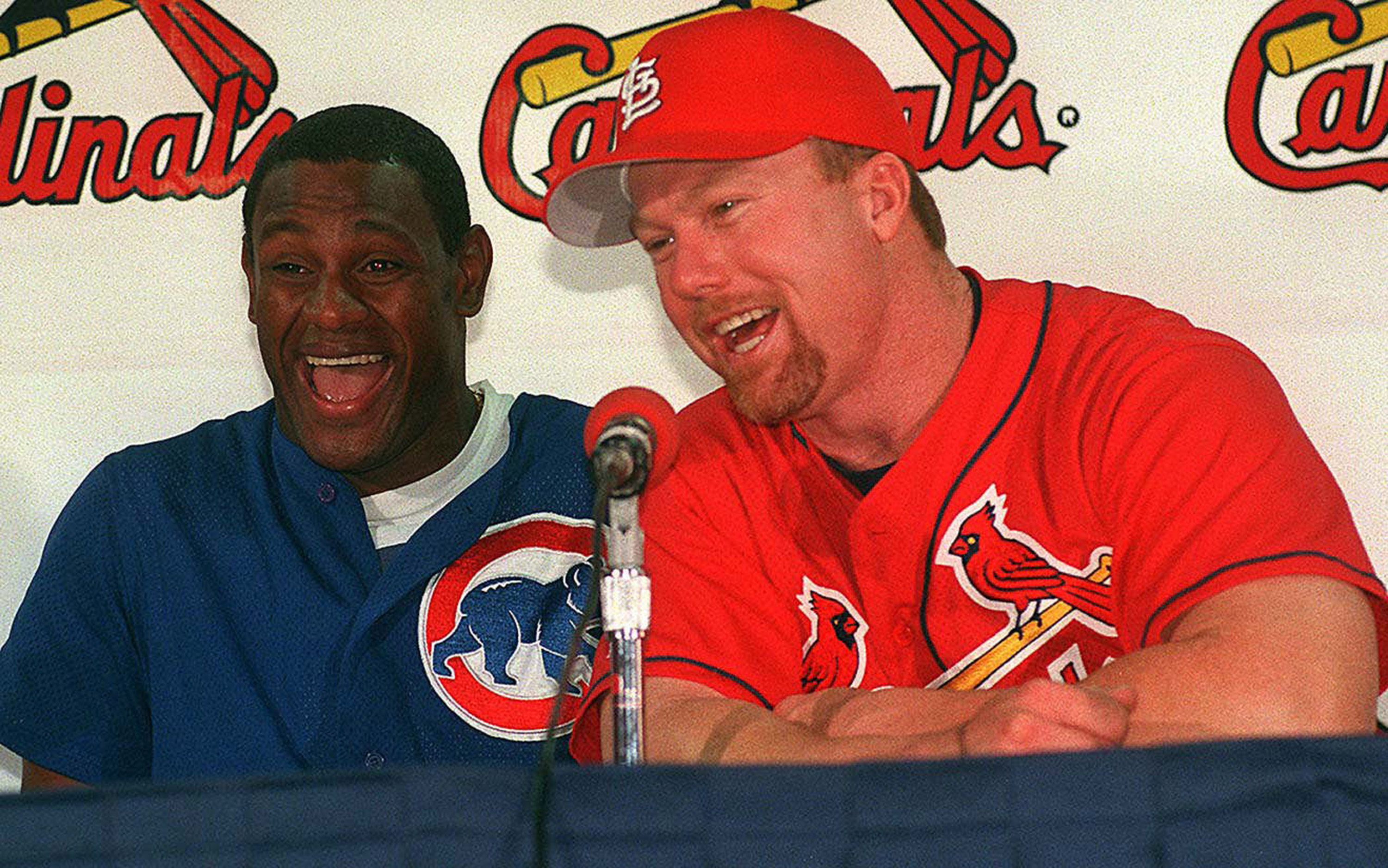 Sammy Sosa and Mark McGwire shared a laugh while speaking with reporters before the Chicago Cubs faced the St. Louis Cardinals on September 8, 1998. Wearing their respective team colors, the two sluggers were locked in a home-run race that had captivated baseball fans nationwide, with both pursuing Roger Maris' long-standing single-season record of 61 as their remarkable seasons unfolded side by side in the national spotlight.