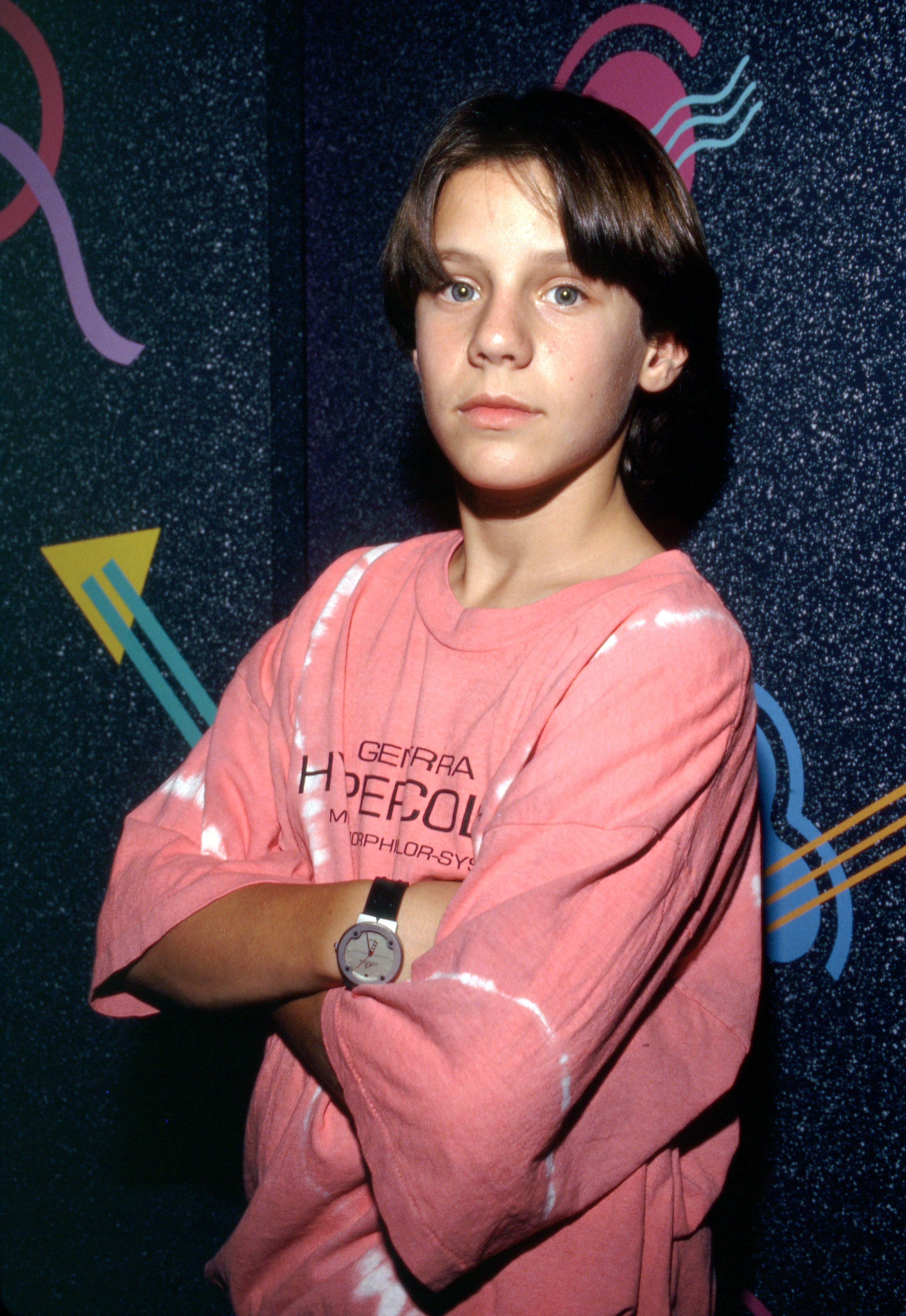 Omri Katz transitions into a cool teen look with longer, center-parted curtain bangs and more mature, angular facial features compared to his early childhood bowl-cut days, wearing an oversized pink Generra Hypercolor t-shirt with his arms crossed. The photo dates back to circa 1990 during the height of his youth stardom.