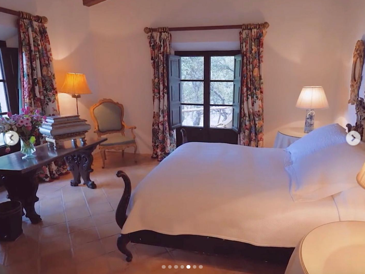 One of the bedrooms takes a quieter approach, with white bedding, floral curtains, warm lamps, and traditional furniture. Windows overlooking the surrounding trees reinforce the sense of privacy that defines much of S'Estaca. | Source: Instagram/robbreport