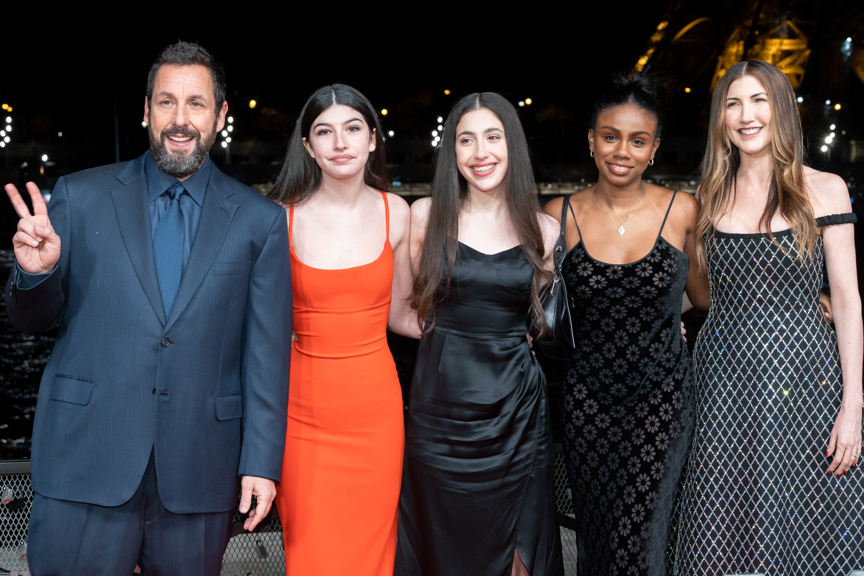 Adam Sandler poses with Sunny, Sadie, Jackie, and a guest against a glowing night backdrop. Sunny wears red, Sadie black satin, and Jackie a patterned gown.