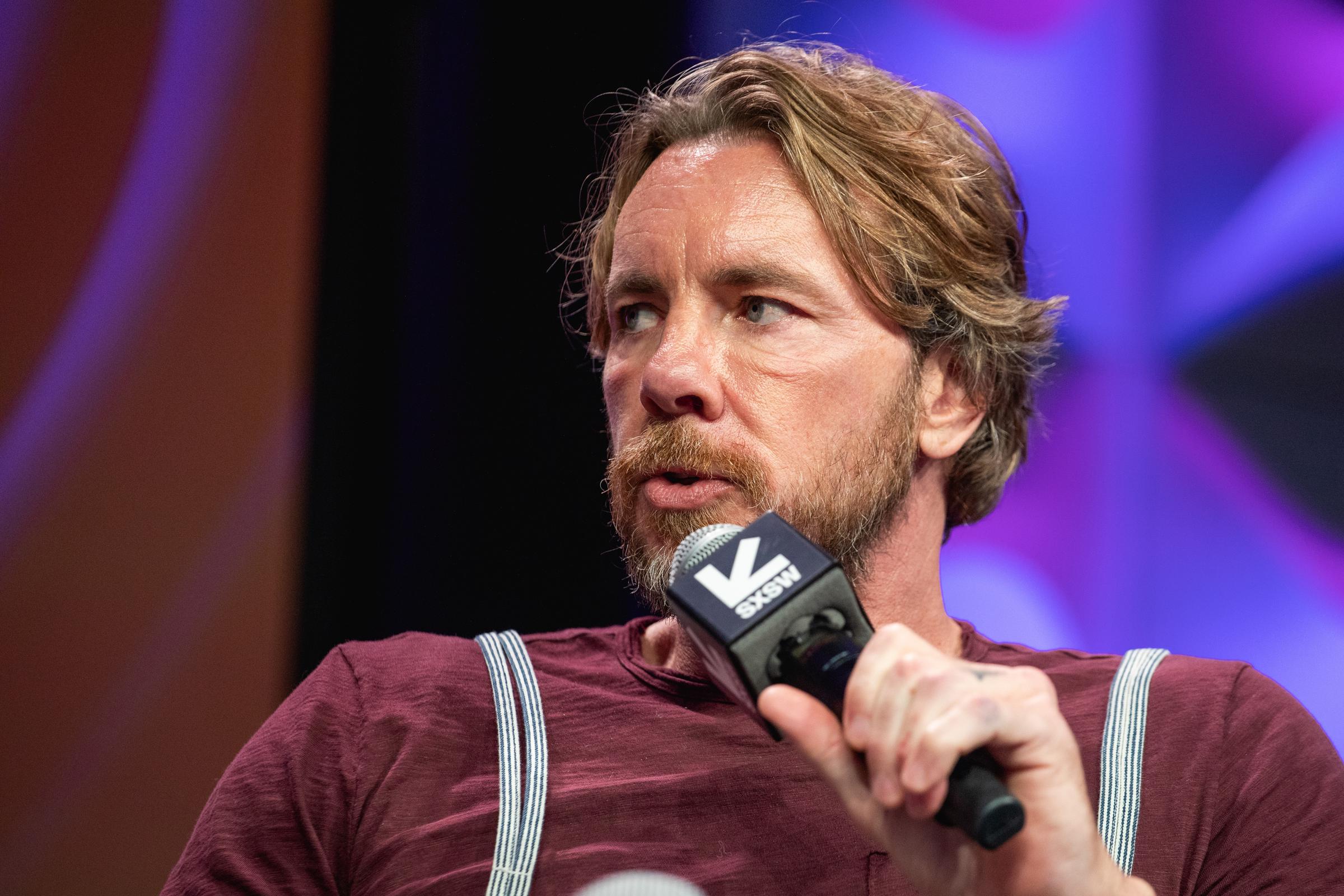 Dax Shepard speaks on stage at SXSW with thoughtful focus during a live session. Wearing a burgundy shirt, he dives into a conversation about connection, creativity, and building a brand through community.