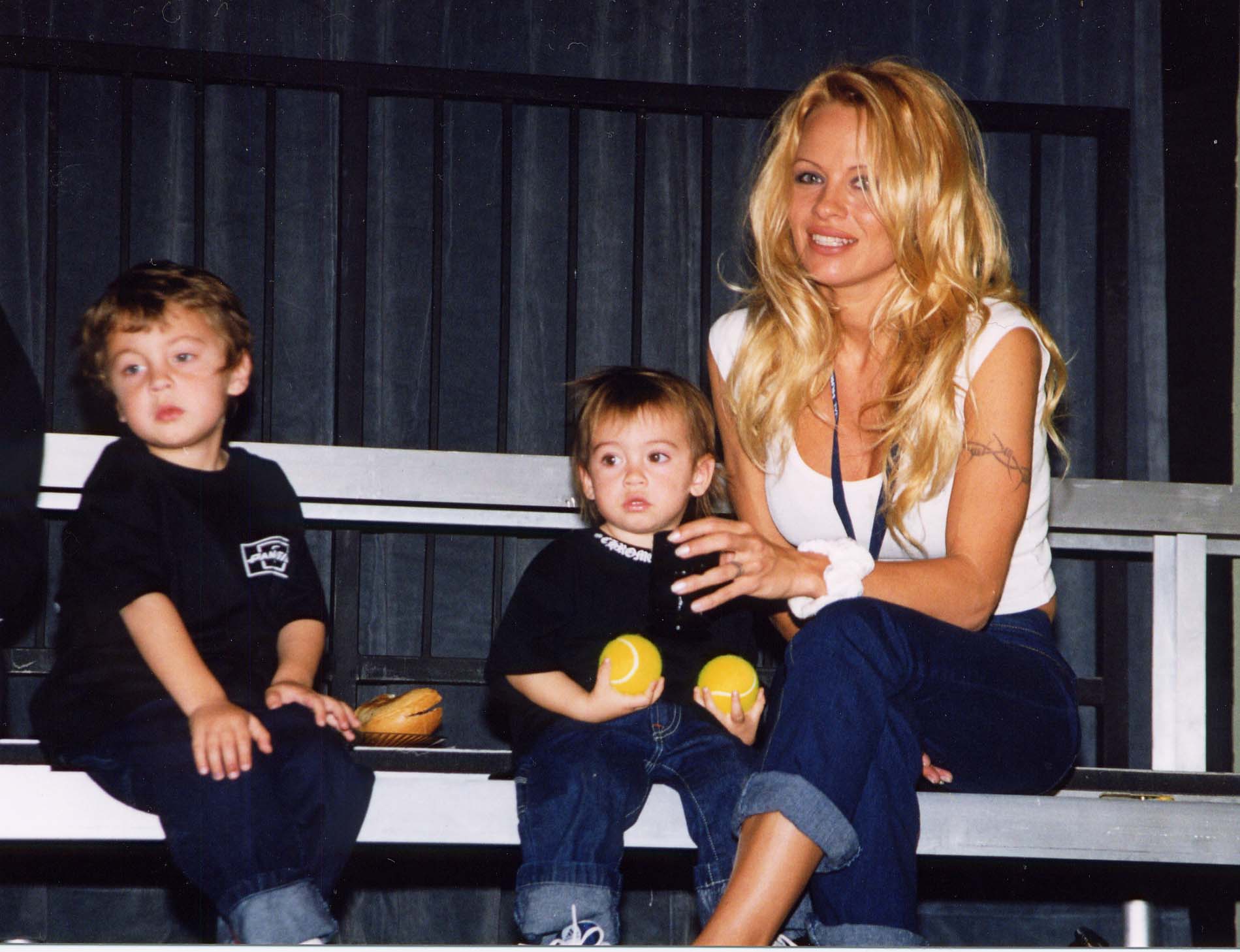 Pamela sits beside her young sons, Brandon and Dylan, during a casual public outing in 1999. Dressed in a white tank top and rolled-up jeans, Anderson smiles warmly while holding Dylan, who clutches two yellow tennis balls. Brandon, seated just beside them in a matching black shirt and jeans, gazes off to the side. The intimate moment, captured on metal bleachers, offers a glimpse into the early years of the Anderson-Lee family.