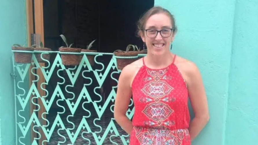 Dr. Julia Pederson smiles warmly in a patterned red dress, standing before a light turquoise wall accented by potted plants along a decorative wrought-iron railing. A beloved pediatrician educated at Harvard and Stanford, she dedicated her life to caring for young children and their families, while quietly cherishing her own 7-year-old daughter before her life was tragically cut short in October 2026. | Source: GoFundMe