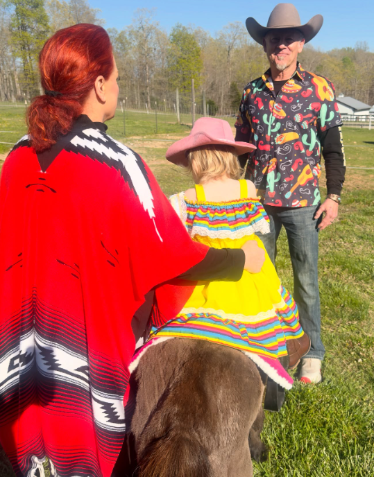 Wearing a bright yellow dress and pink cowboy hat, Kaliyah sits on a small pony while Wynonna stands beside her and Cactus smiles nearby. The photograph brings together three generations of the family in a grassy outdoor setting. | Source: Instagram/wynonnajudd