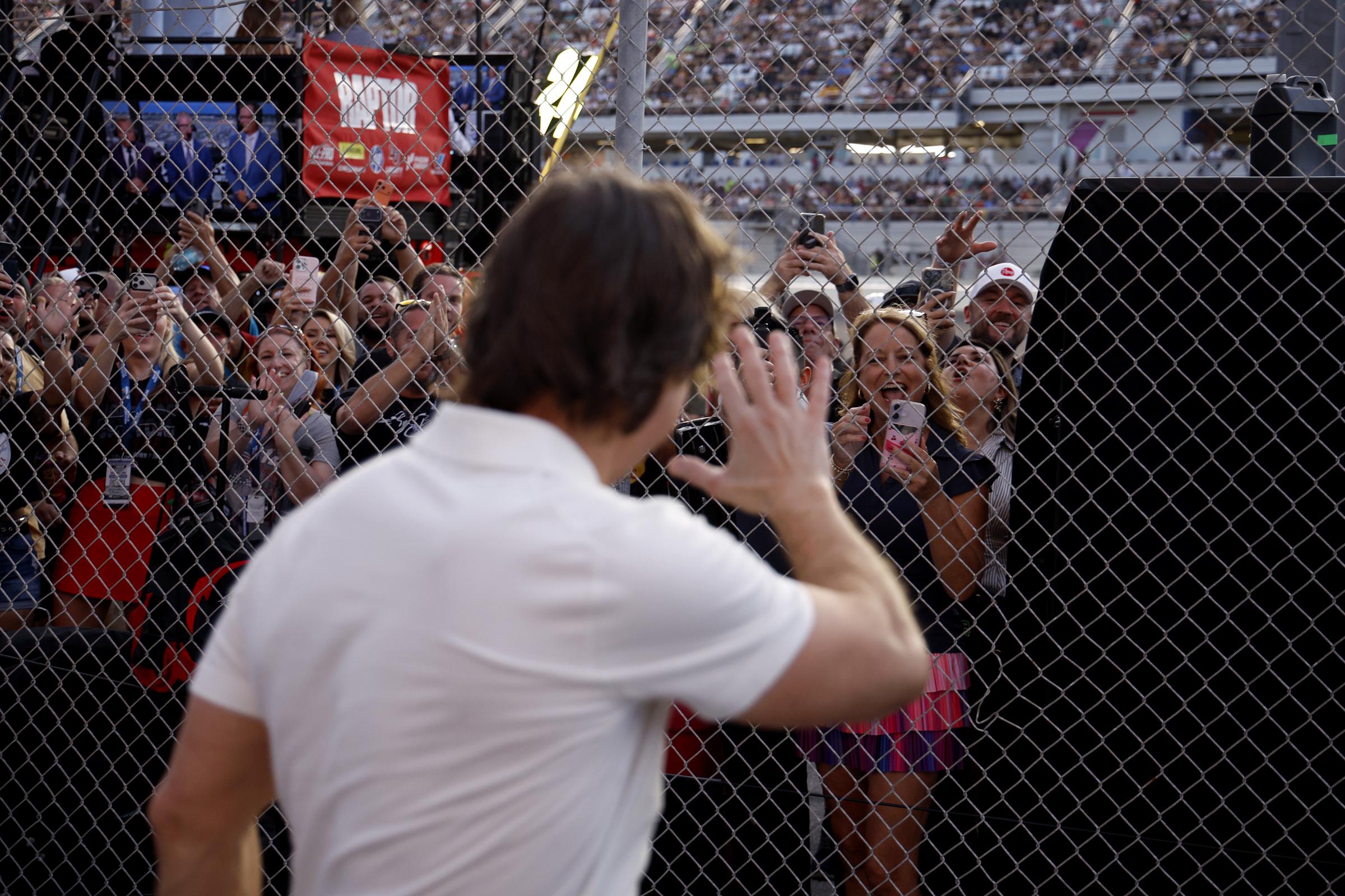 Cruise waves to fans gathered behind a fence at Daytona International Speedway as several people raise their phones to capture the moment. The actor is seen from behind in his white polo, with the crowded speedway visible beyond the fence. | Source: Getty Images