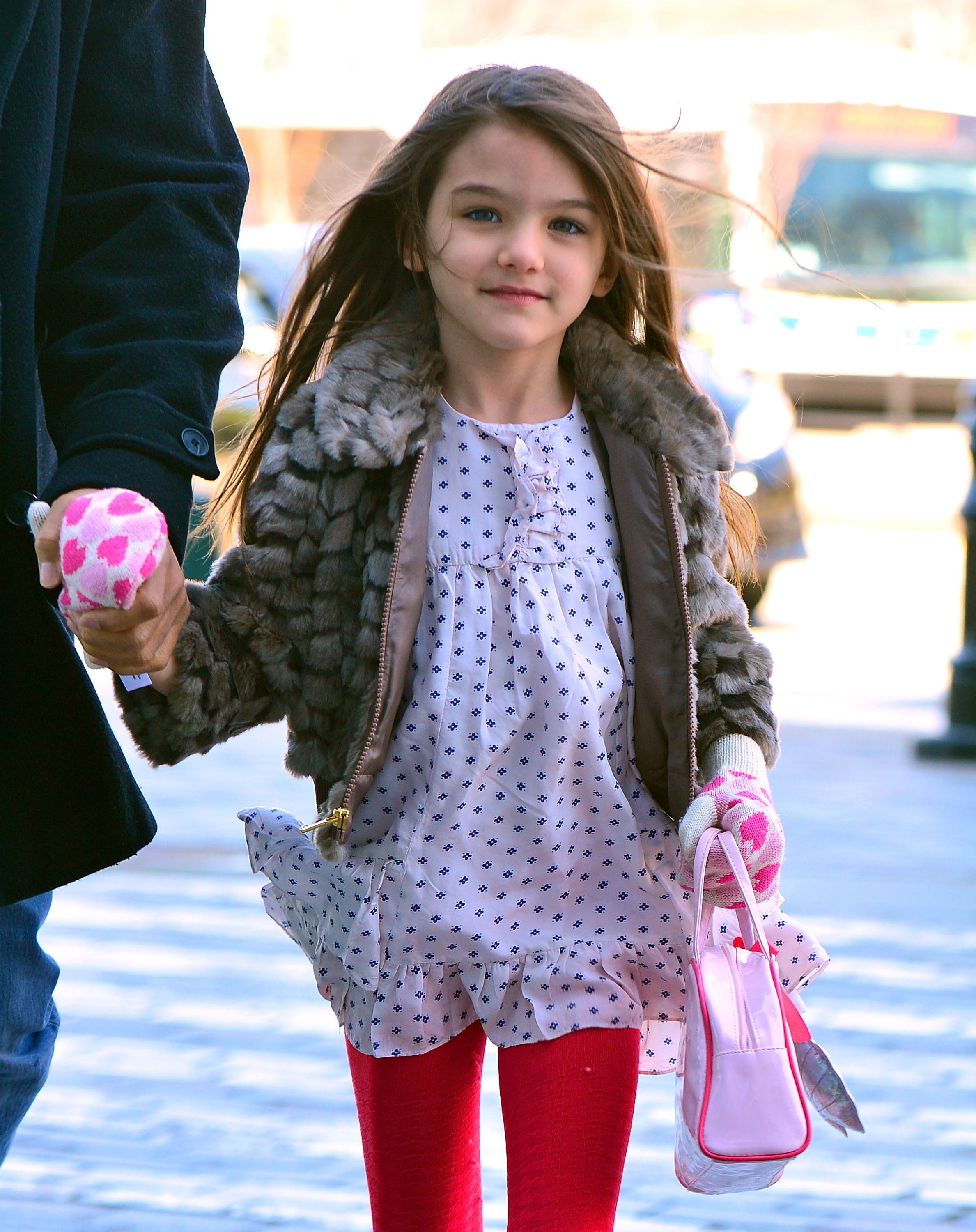 Dressed in a playful polka dot dress and bright red leggings, little Suri Cruise is captured mid-stroll, her small hand clasped securely in another’s. Her charm shines through as she walks down the street, a picture of childhood innocence amid the flash of paparazzi lenses.