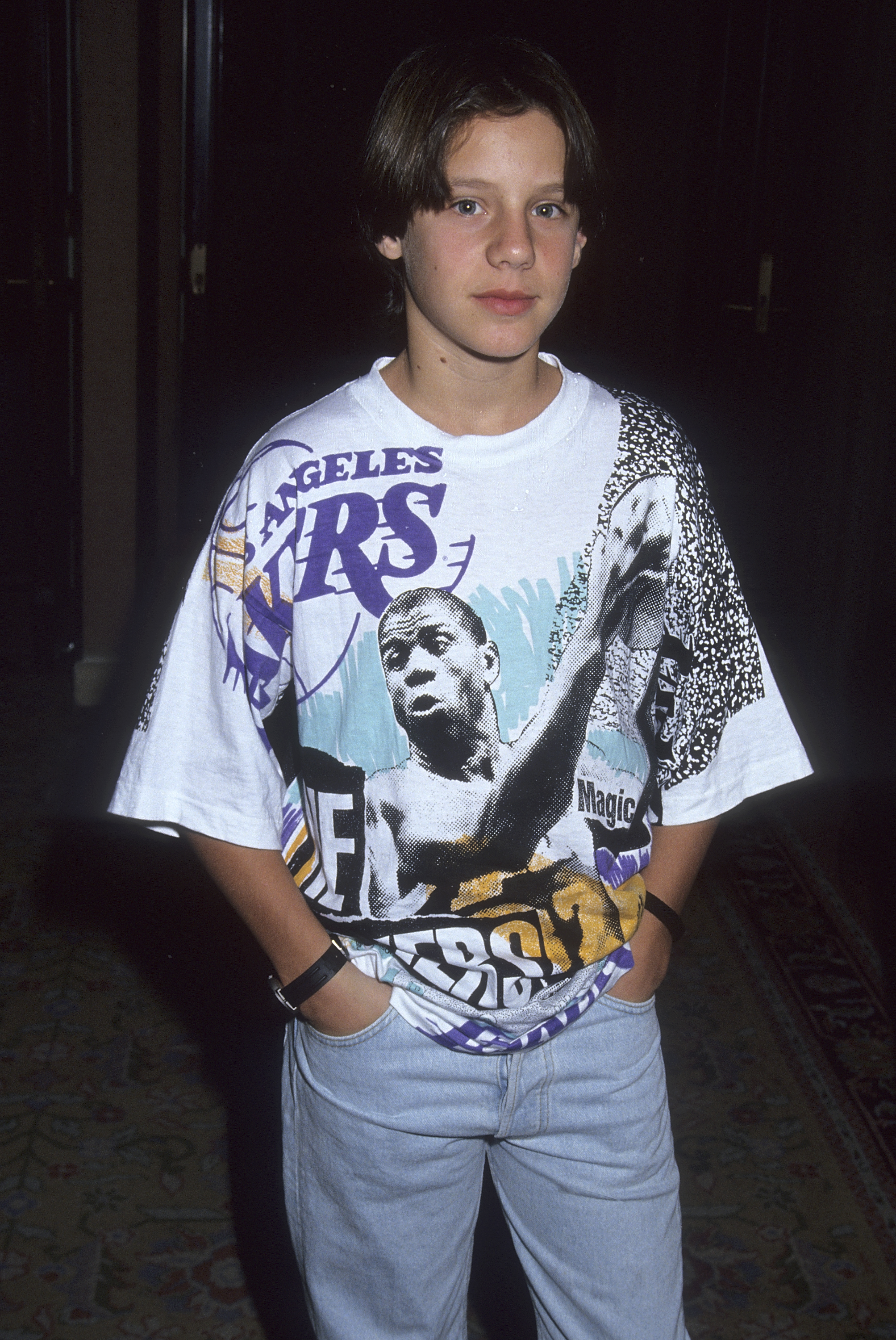 Omri Katz continues to grow into his teen years with a taller, leaner frame and chin-grazing dark curtain bangs that replace his early childhood neat cuts, wearing a graphic oversized Magic Johnson Los Angeles Lakers t-shirt paired with light-wash denim jeans. The photo was taken on July 27, 1991, as he attended the NBC Summer TCA Press Tour at the Universal Hilton Hotel in Universal City, California.