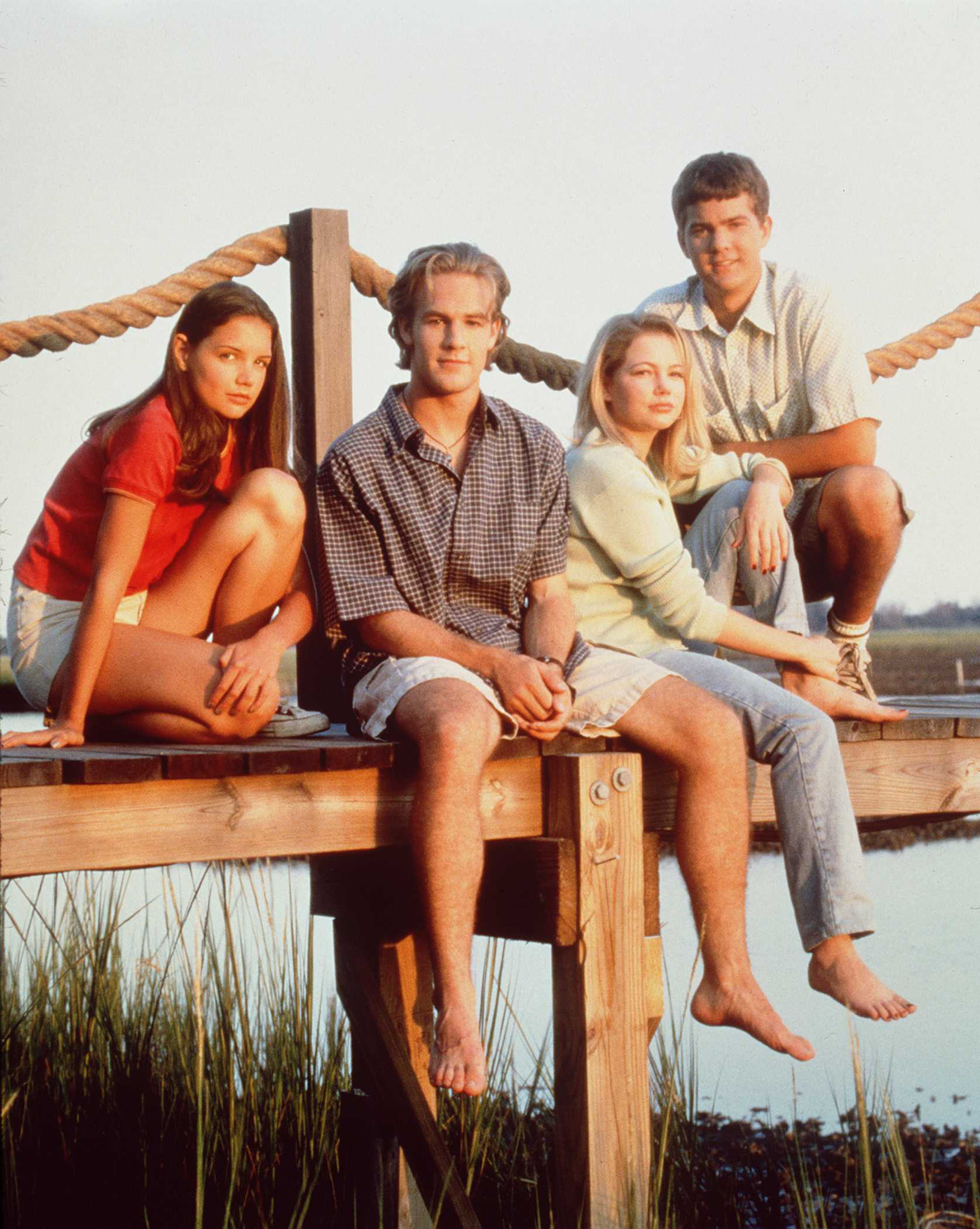 Katie Holmes, James Van Der Beek, Michelle Williams, and Joshua Jackson sit closely on a wooden dock beside calm water, bathed in warm, soft light. The scene captures a relaxed, summery moment against a peaceful outdoor backdrop.