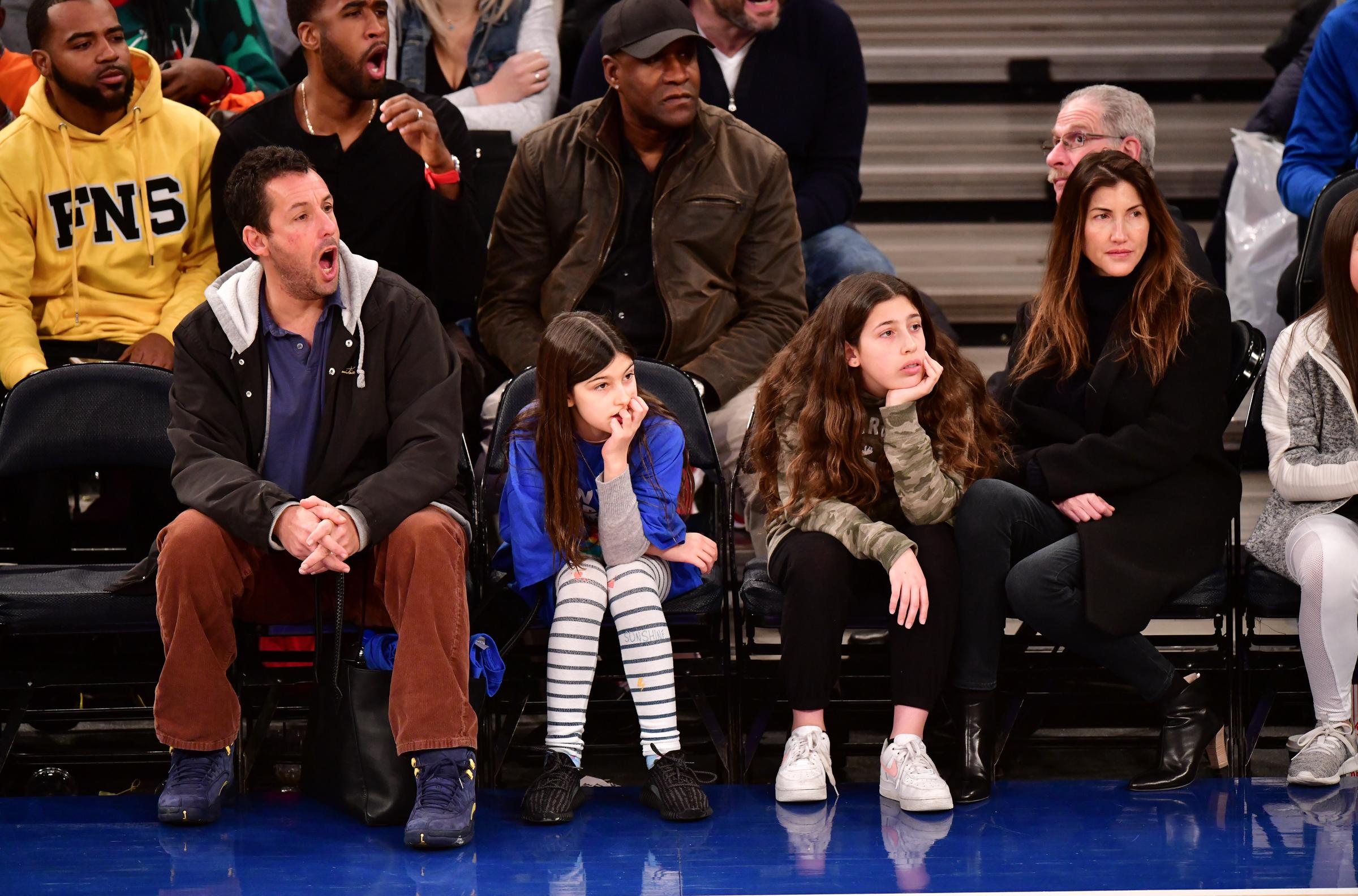 Adam Sandler reacts mid-play with an open-mouthed cheer as Sunny and Sadie sit beside him, while Jackie Sandler watches calmly from the end of the row. From striped leggings to cozy layers, the family’s relaxed courtside style contrasts with the intensity of the Christmas Day matchup.