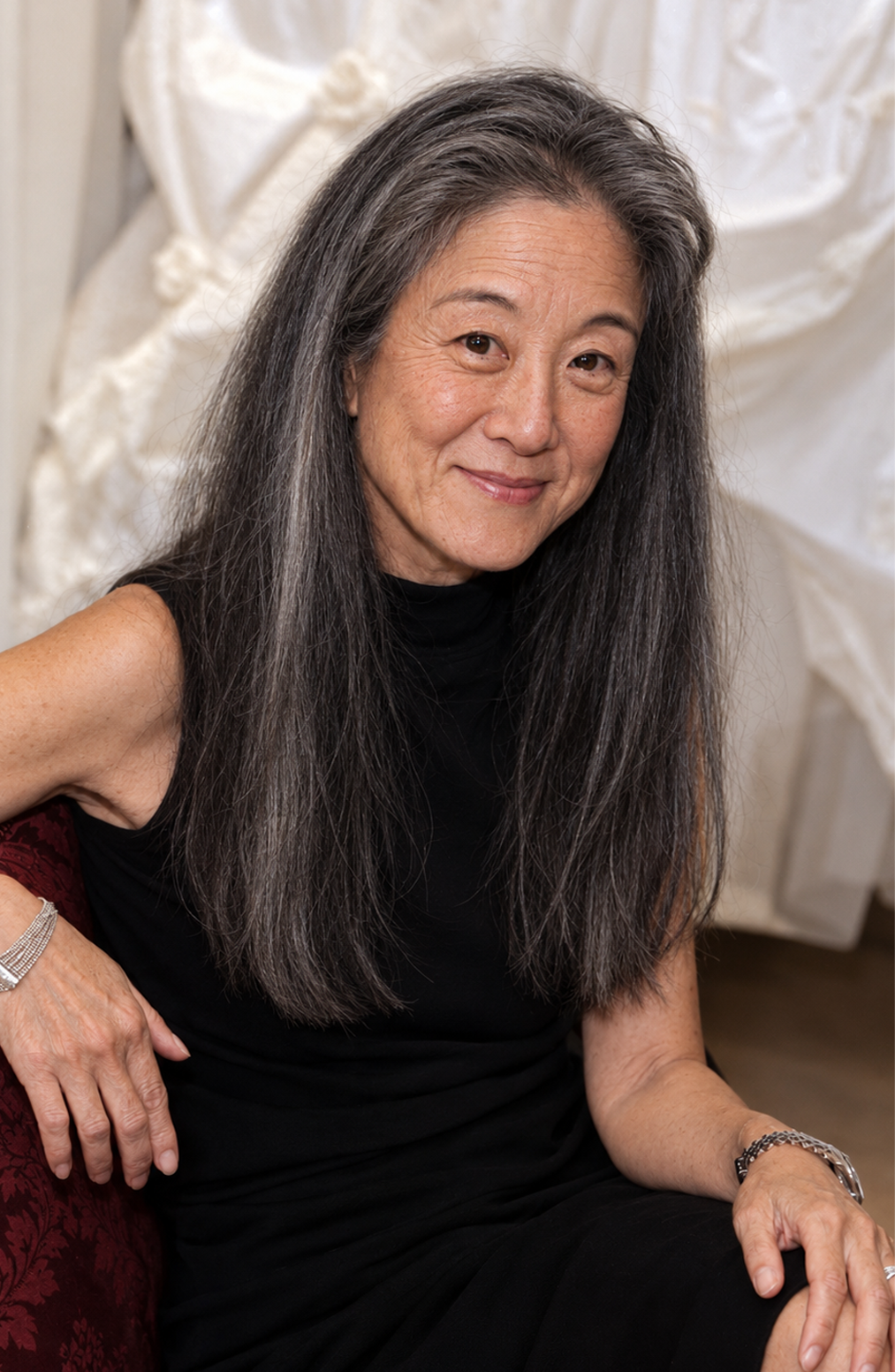 Revisiting her iconic 1990 bridal debut photoshoot through an older lens, this AI concept portrays Vera Wang seated on a settee before draped white gowns with natural salt-and-pepper tresses and subtle character lines. The digital reimagining emphasizes her original facial contours and wider nose bridge, accented with twin silver cuffs and a sleeveless black dress. | Source: ChatGPT