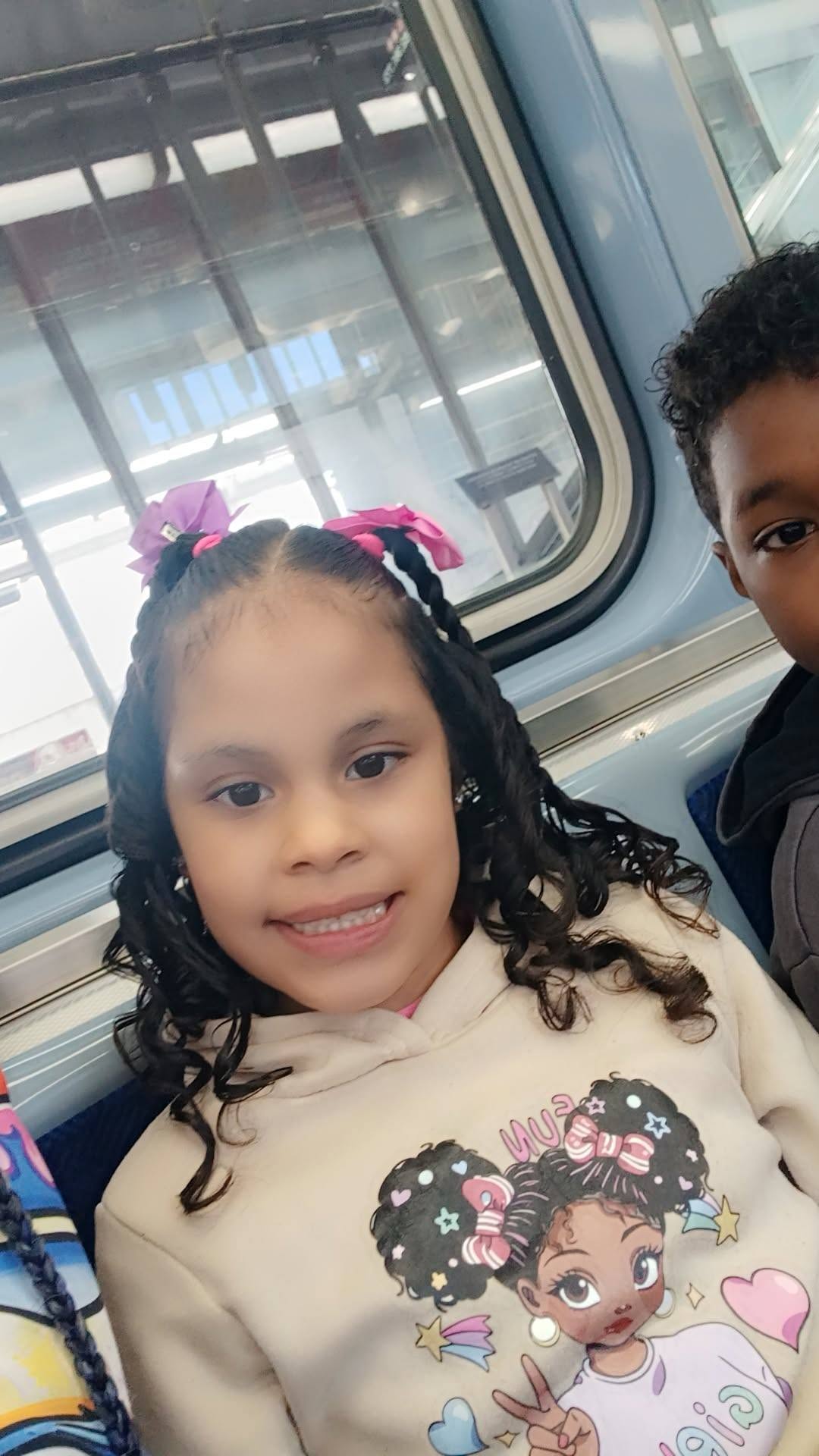 Eddimar Paola Hernández Chacón flashed a sweet smile while sitting on a subway train with her brother visible beside her. Her hair was styled in curly braids accented with bright pink and purple bows, and she wore a cream-colored hoodie featuring a colorful cartoon girl graphic. | Source: Facebook/Morena Esa