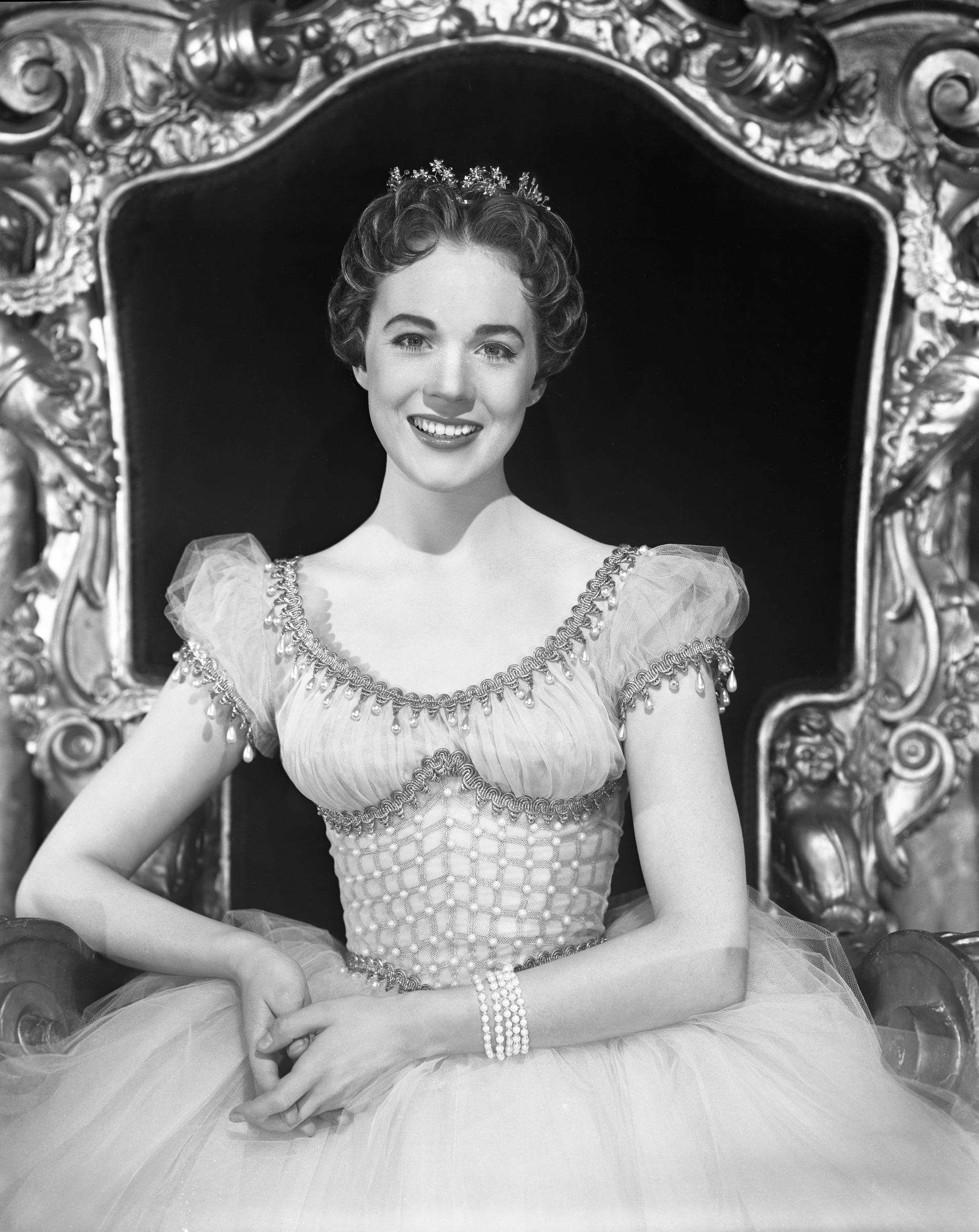 Julie Andrews embodies fairytale royalty in a promotional portrait for the 1957 CBS television musical "Cinderella," written by Richard Rodgers and Oscar Hammerstein II, photographed on January 8, 1957, in New York City. Seated gracefully before an ornate, carved royal throne, the actress beams in an off-the-shoulder tulle ballgown accented with pearl latticework, teardrop beading along the neckline, a multi-strand pearl bracelet, and a delicate floral tiara in her curled hair.