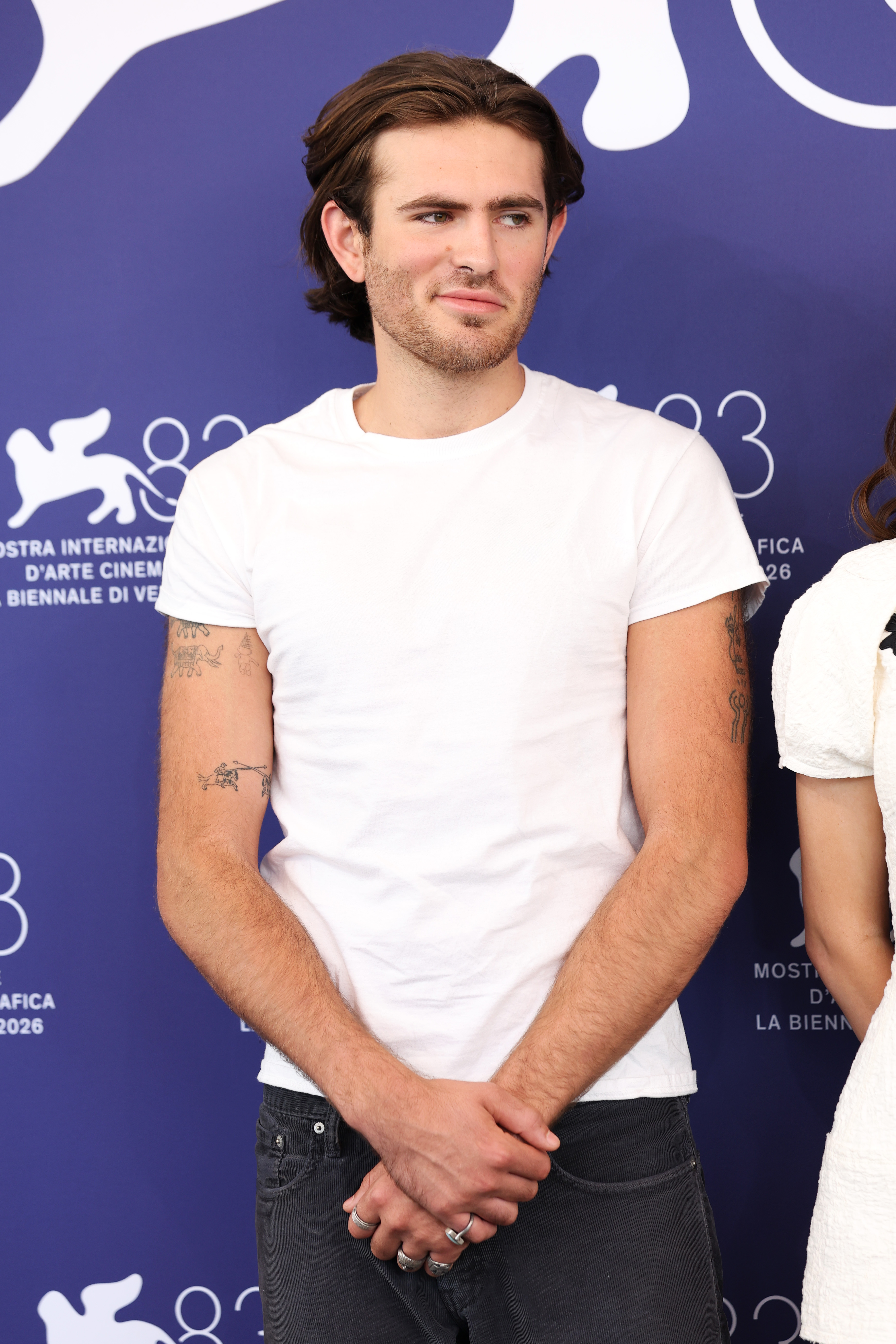 Opting for an understated, casual daytime look, Indiana Affleck posed in a simple white crewneck T-shirt and charcoal denim at the 83rd Venice International Film Festival photocall on September 8, 2026. The short-sleeved cut put a collection of delicate, fine-line arm tattoos on display, while stacked silver rings and a subtle, knowing smirk added a laid-back edge against the vibrant festival backdrop. | Source: Getty Images