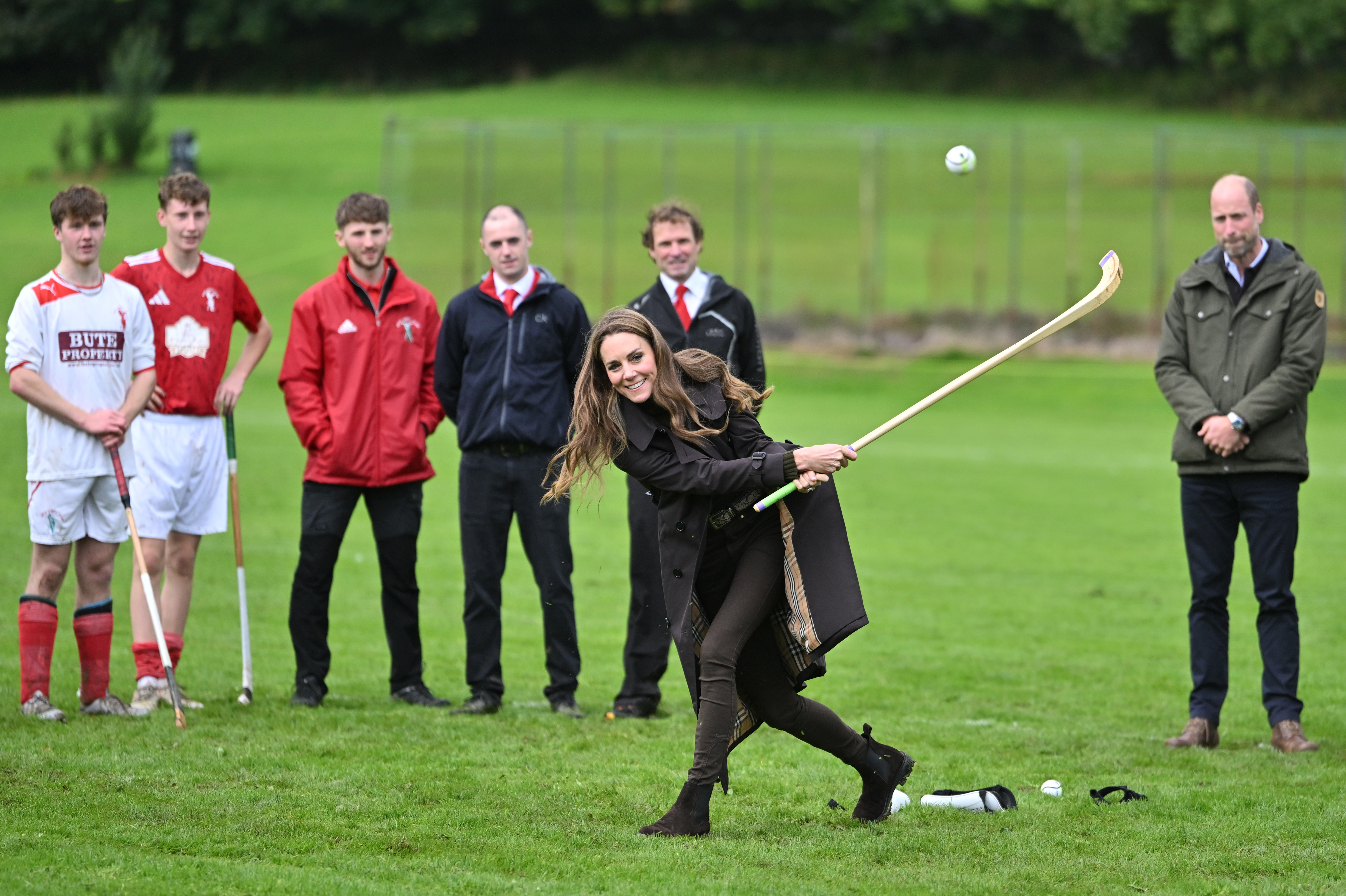 Catherine, Princess of Wales, follows through on her swing as the shinty ball flies across the pitch, with Prince William and players watching the attempt from behind. | Source: Getty Images