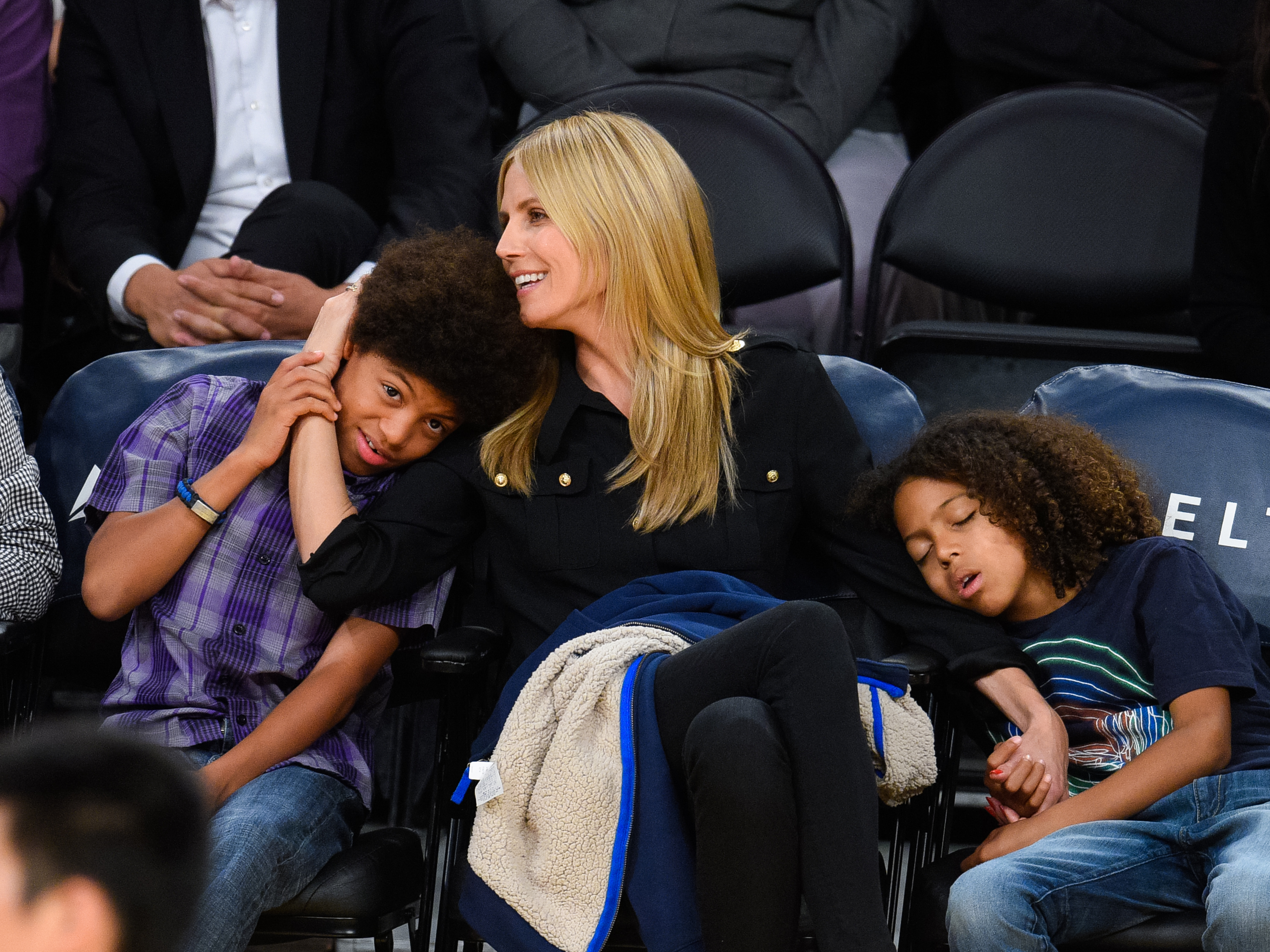 Heidi Klum shared a tender courtside moment with her sons, affectionately cuddling one while the other slept peacefully on the chair beside her. Dressed casually, the family radiated warmth and closeness amid the lively sports arena backdrop.