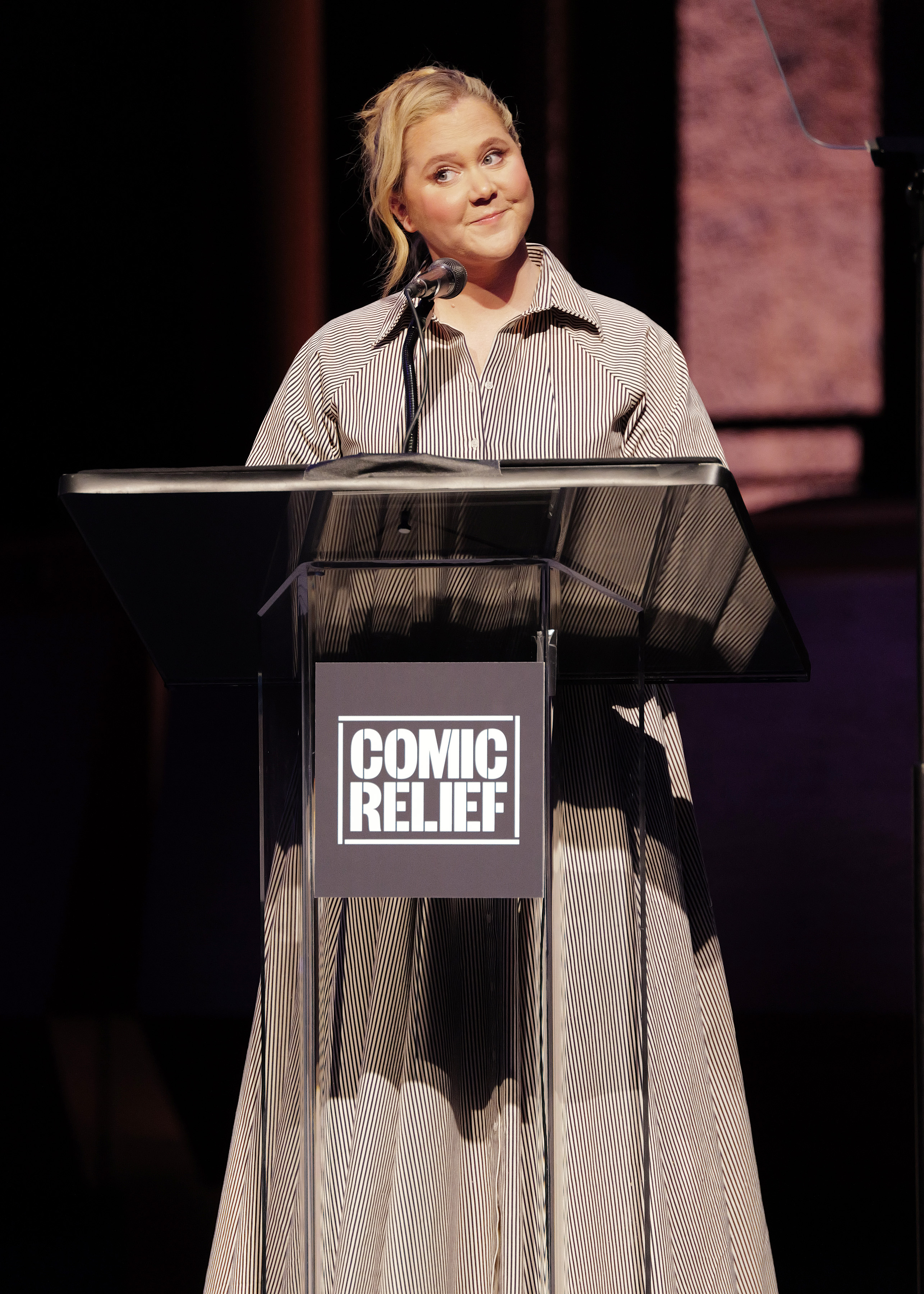 Amy Schumer stands at the podium while speaking during Comic Relief’s Icon Award event in New York City. She wears a long striped dress with a collared neckline and her hair pulled back in a loose updo.