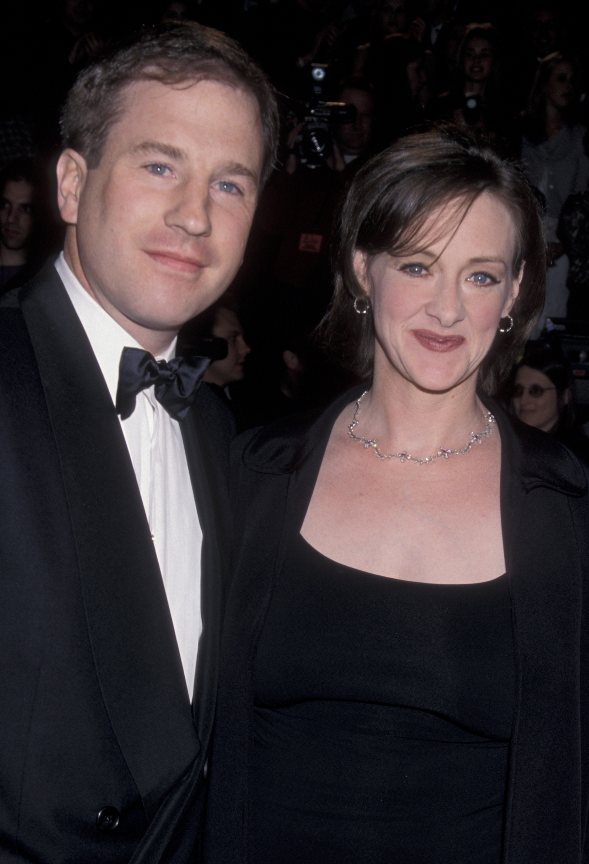 Joan Cusack and Richard Burke pose together in coordinated black formalwear at a glamorous event. Her sparkling necklace and warm smile add a touch of refinement to the couple's look.