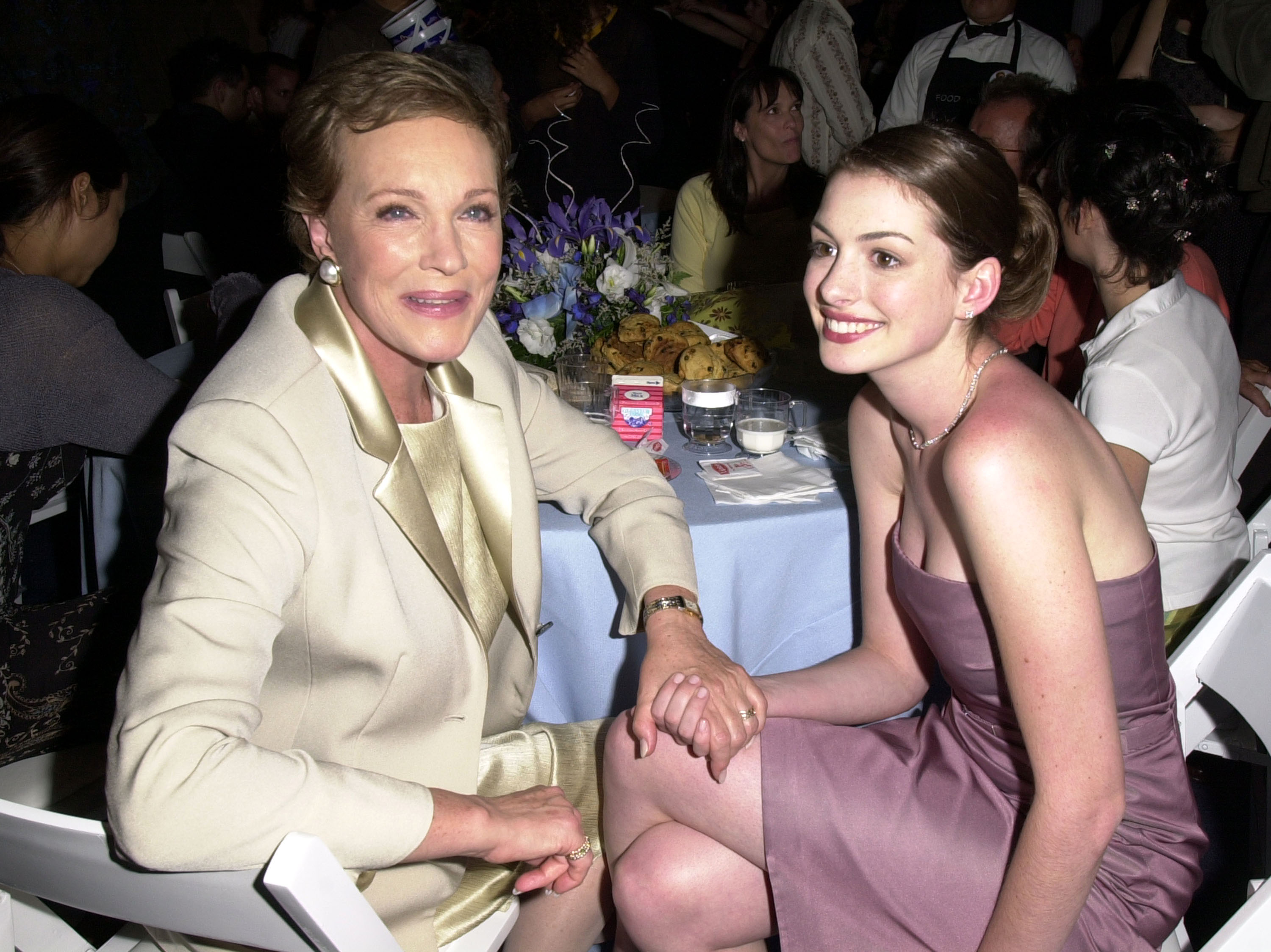 Julie Andrews and a young Anne Hathaway share an affectionate, smiling moment seated together at the premiere after-party for "The Princess Diaries" at the El Capitan Theatre in Hollywood, California, on July 29, 2001. Embodying their regal on-screen bond, Andrews holds Hathaway's hand while wearing an elegant champagne-toned tailored blazer and matching satin top, paired with classic pearl earrings, while Hathaway shines in a strapless mauve cocktail dress and a dainty necklace.