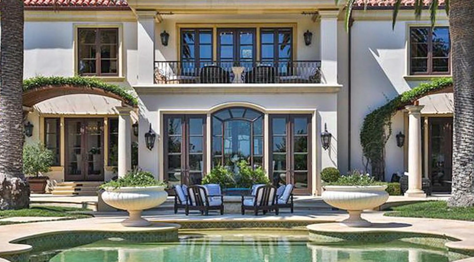 The rear facade of the Beverly Hills mansion opens toward a tranquil Grecian-style swimming pool, framed by stone urn planters, arched ivy-draped pergolas, and French doors leading out from the main level beneath a second-story balustraded balcony. | Source: YouTube/Taste of Country
