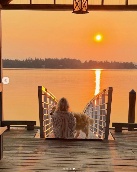 Pamela Anderson embraces the peaceful rhythms of coastal life at Arcady, sitting beside one of her dogs on a wooden pier during a radiant sunset in Ladysmith, British Columbia. Viewed from behind in an oversized striped button-down, she looks out across the glowing water as the evening sun dips behind the treeline, sharing the serene moment with followers alongside the caption, "An island girl at heart 🤍." | Source: Instagram/pamelaanderson