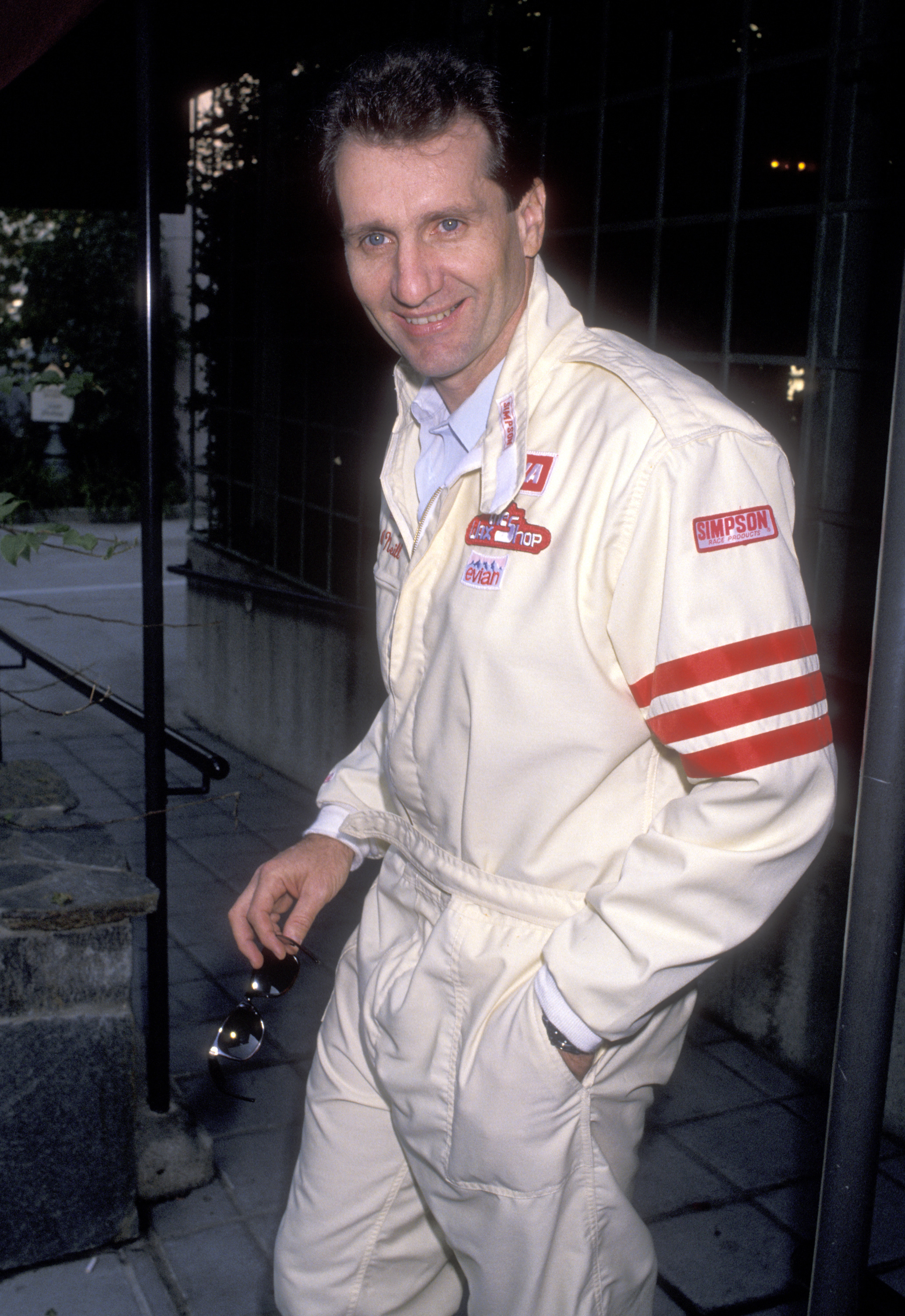 Ed O'Neill suited up in a cream racing jumpsuit complete with red arm stripes and sponsor patches while attending the Great American Road Rally Benefit Second Chance And Bread For Life on March 11, 1989, at Ma Maison Sofitel in Los Angeles, California. Holding a pair of sunglasses with one hand casually tucked in his pocket, the "Married... with Children" star smiled for photographers outside the venue before taking part in the charity event. | Source: Getty Images