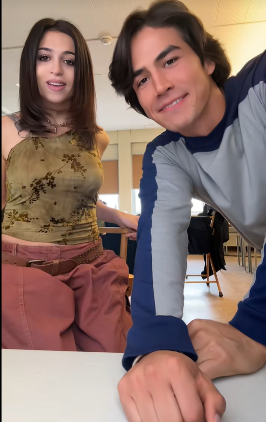 Josie Totah appears in a TikTok video with a male companion, sitting beside him at a table in what appears to be a rehearsal or studio space. She wears an olive-green floral top, dusty-rose cargo pants, and layered necklaces, while her companion wears a gray-and-navy long-sleeved shirt. | Source: TikTok/josie totah