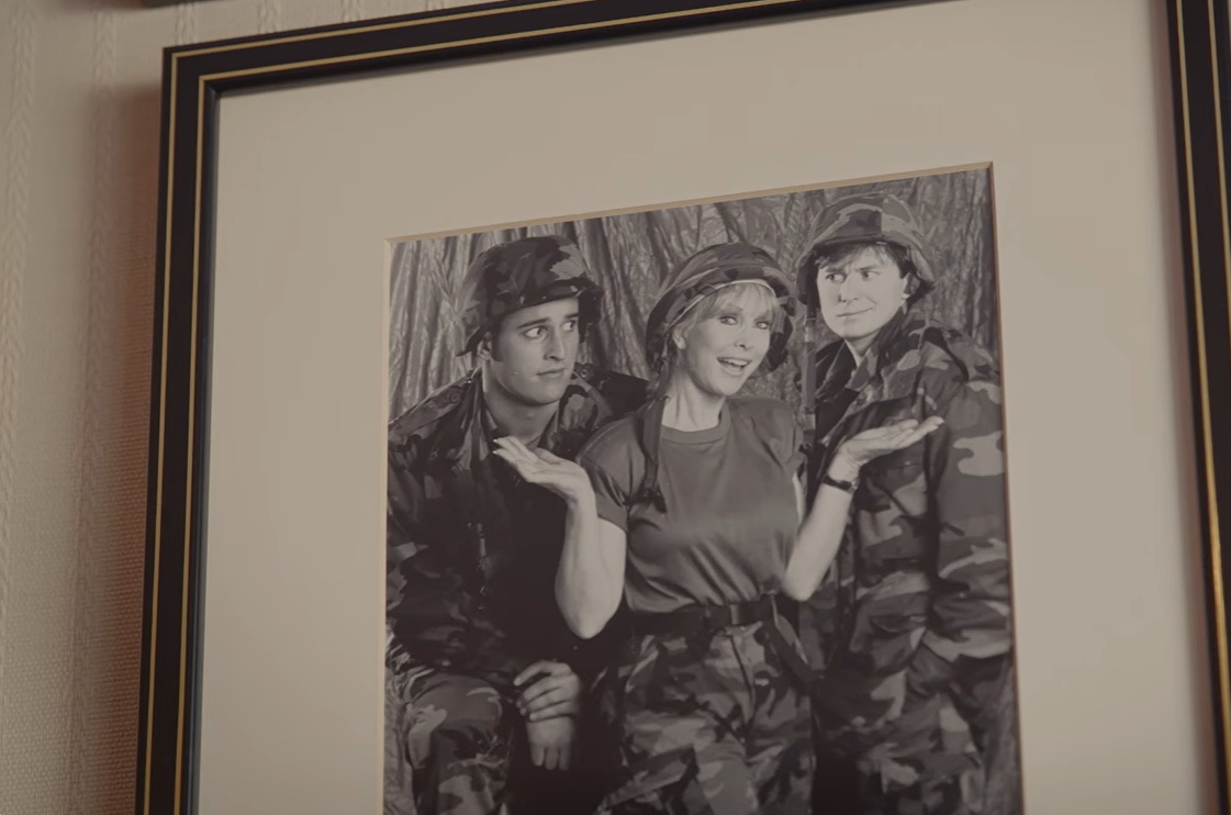 A framed black-and-white photograph shows Barbara Eden smiling between her son, Matthew Ansara, and actor Paul Eiding in military-style costumes for the television movie Your Mother Wears Combat Boots. | Source: YouTube/People