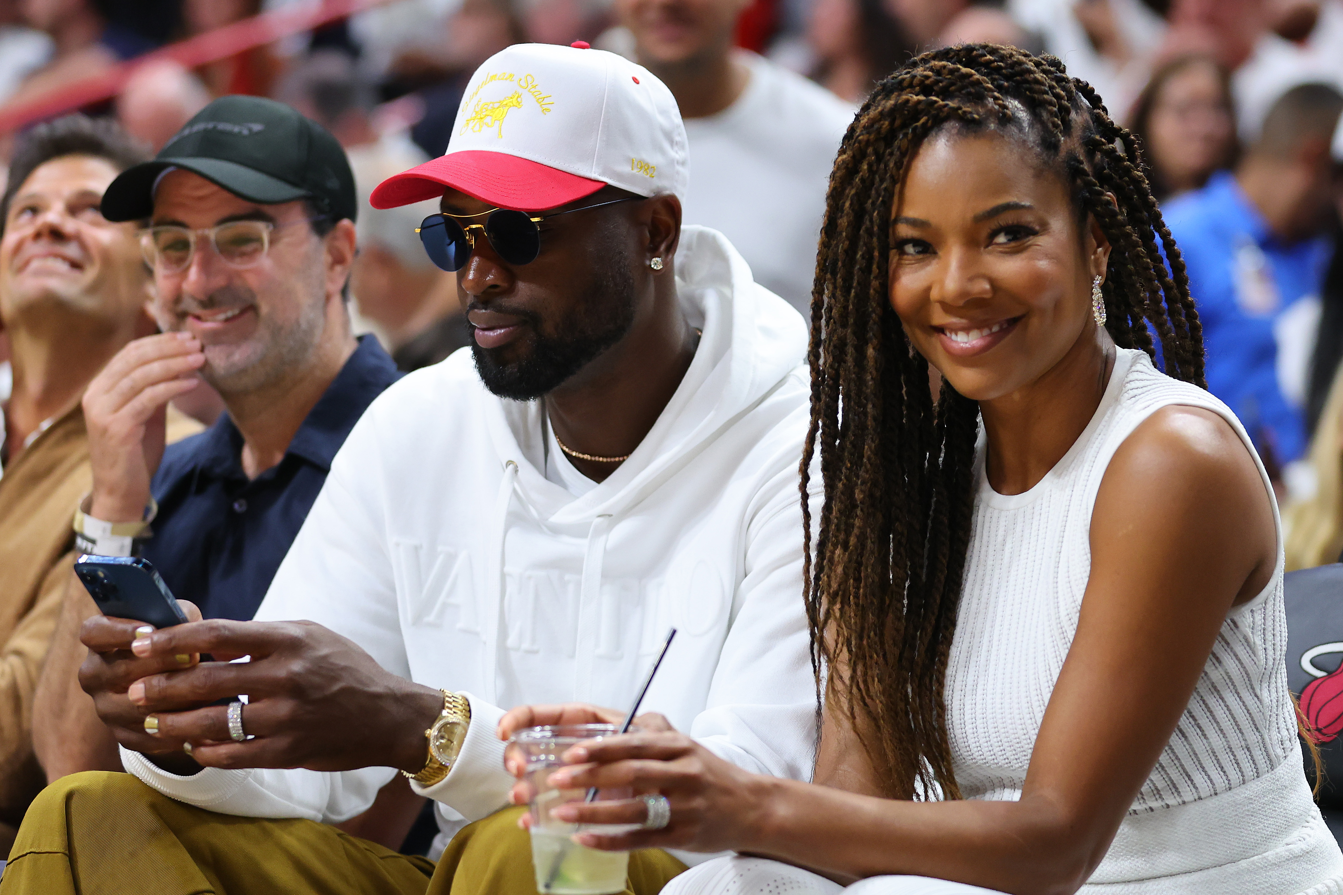 Gabrielle Union sits courtside beside Dwyane Wade at a basketball game. She wears a sleeveless white top and long braids, smiling toward the camera while holding a drink. | Source: Getty Images