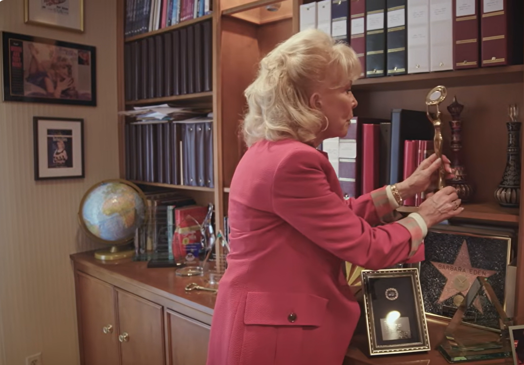 Barbara Eden carefully adjusts a gold-colored trophy on her office shelf, where a framed Hollywood Walk of Fame star bearing her name sits among other awards, plaques, and career memorabilia. | Source: YouTube/People