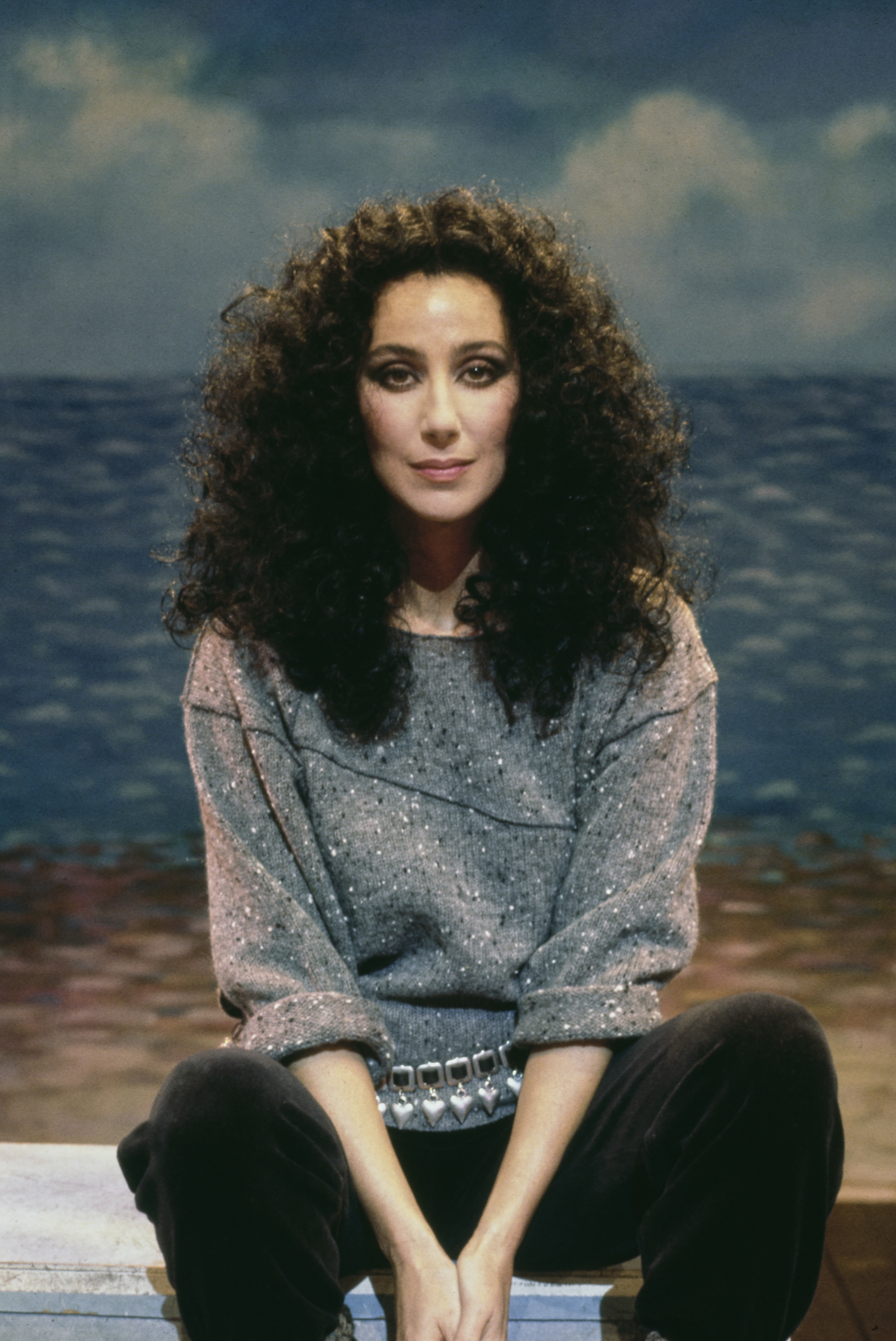 Music and cultural icon Cher sat cross-legged with her hands resting between her knees in front of a painted beach backdrop in 1983. She framed her gaze with voluminous, cascading dark curls, dressed casually in a grey flecked knit sweater with rolled sleeves, accessorized with a metallic heart-charm belt over dark trousers. | Source: Getty Images