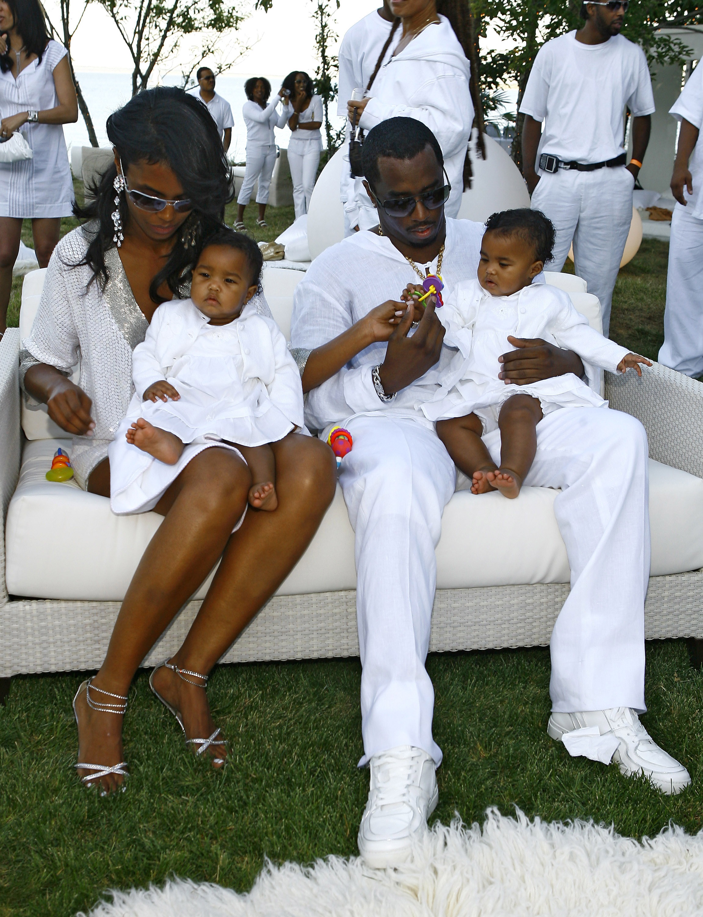 Sean “Diddy” Combs and Kim Porter sit together with their twin daughters, D’Lila Star and Jessie James Combs, all dressed in white as they hold the girls during a sunlit outdoor summer gathering.