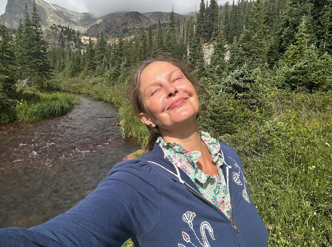 Judd closes her eyes and smiles in the sunshine during a 2025 mountain outing. Wearing a blue hoodie beside a stream, she shares a relaxed moment far from the red carpet. | Source: Facebook/AshleyJuddOfficial