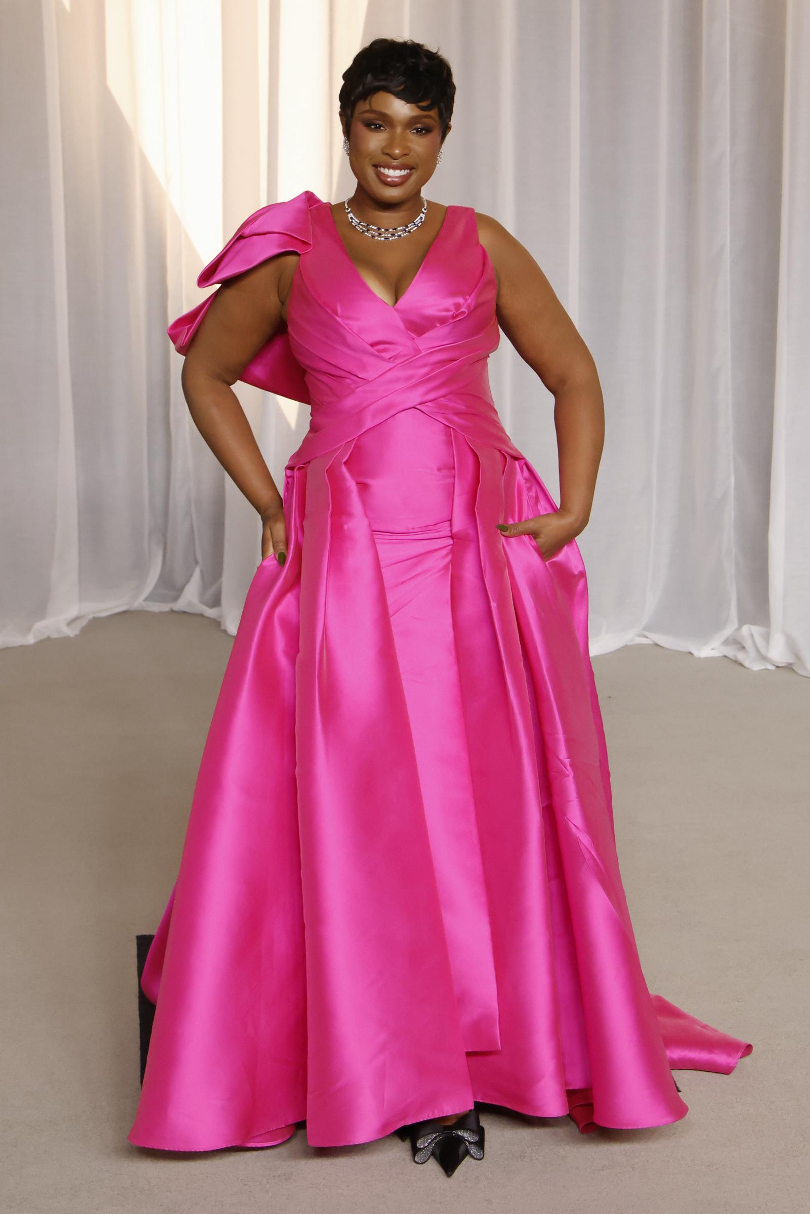 Jennifer Hudson commands attention in a vibrant, hot pink satin gown featuring a plunging V-neckline and sculpted drapery along the bodice. An asymmetrical, voluminous bow accents one shoulder, while the full, floor-sweeping skirt includes built-in pockets for effortless charm. The EGOT winner completes the showstopping look with a sparkling diamond collar necklace, pointed-toe heels adorned with crystal bows, and a sleek pixie cut.