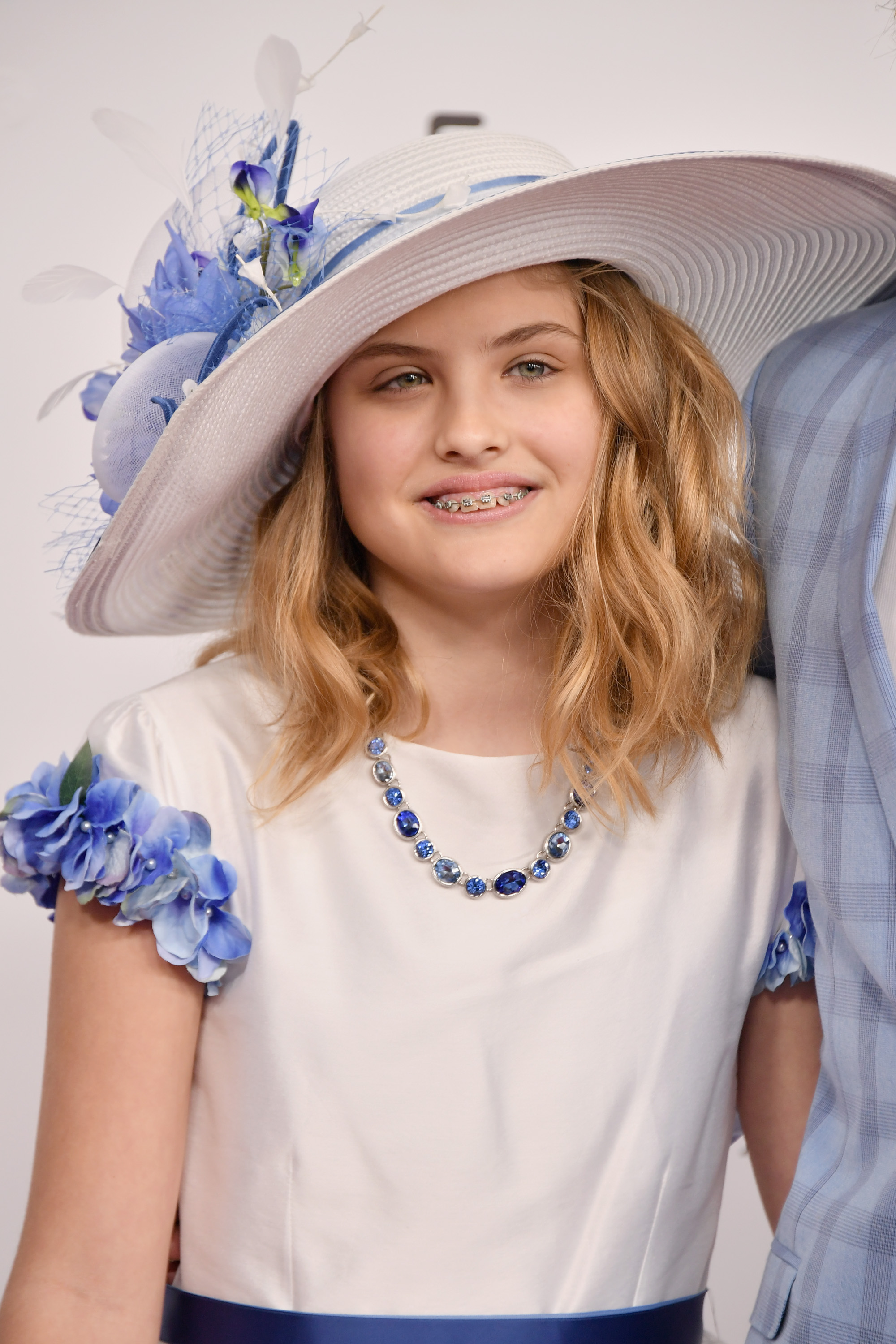 A broad white sun hat styled with voluminous blue feathers and fine mesh netting framed 11-year-old Dannielynn Birkhead at the 144th Kentucky Derby on May 5, 2018. Outfitted in a pristine white dress accented by vibrant blue floral sleeve cuffs and a matching jewel-toned necklace, she offered a bright, brace-adorned smile that captured a transitioning milestone into pre-teen independence.