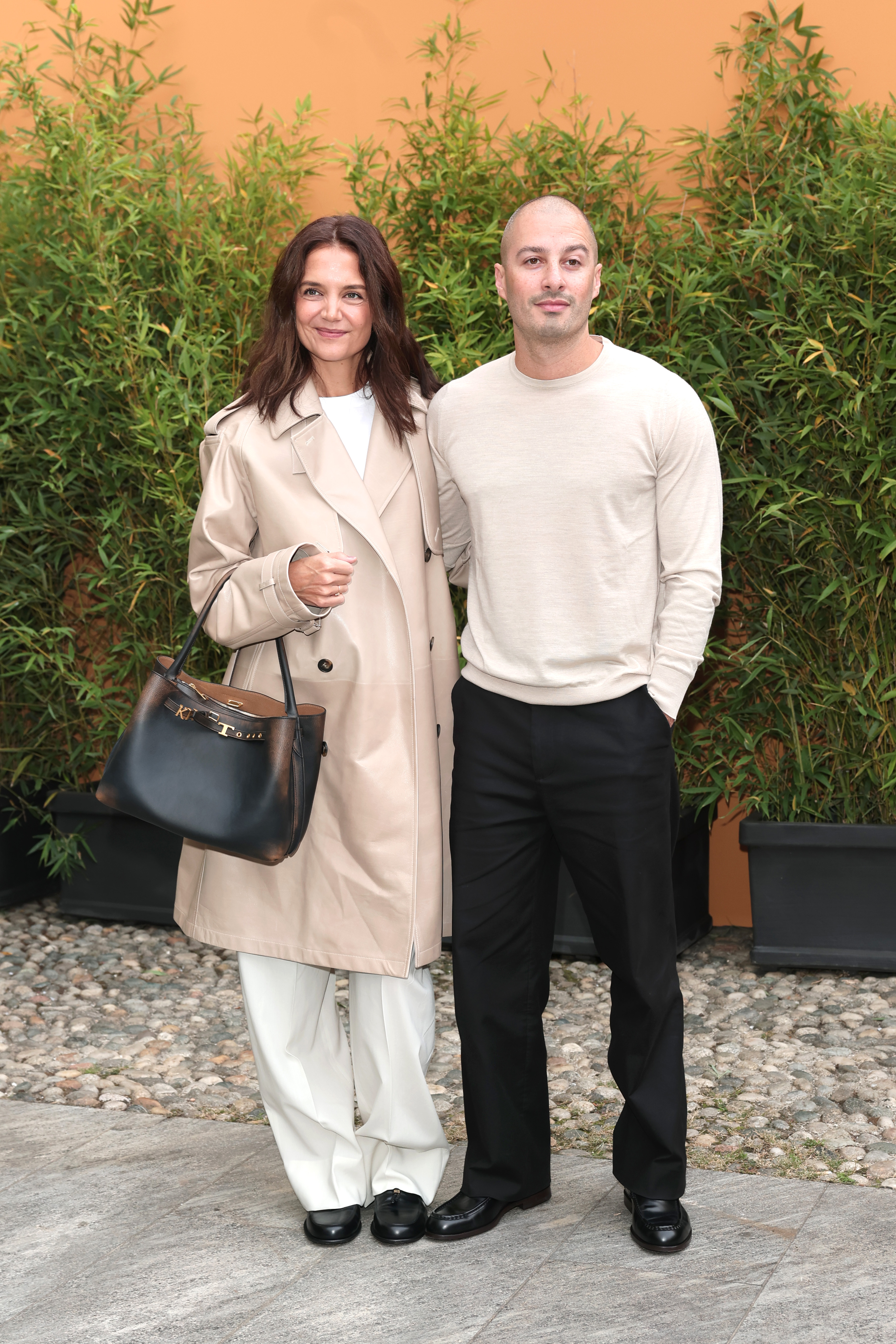 Standing against a wall of lush potted bamboo, the couple paused for a serene official portrait before heading inside to take their seats on September 25, 2026, in Milan. Katie flashed a soft, content smile directly toward the cameras while Jason stood at ease with one hand resting in his pocket. | Source: Getty Images