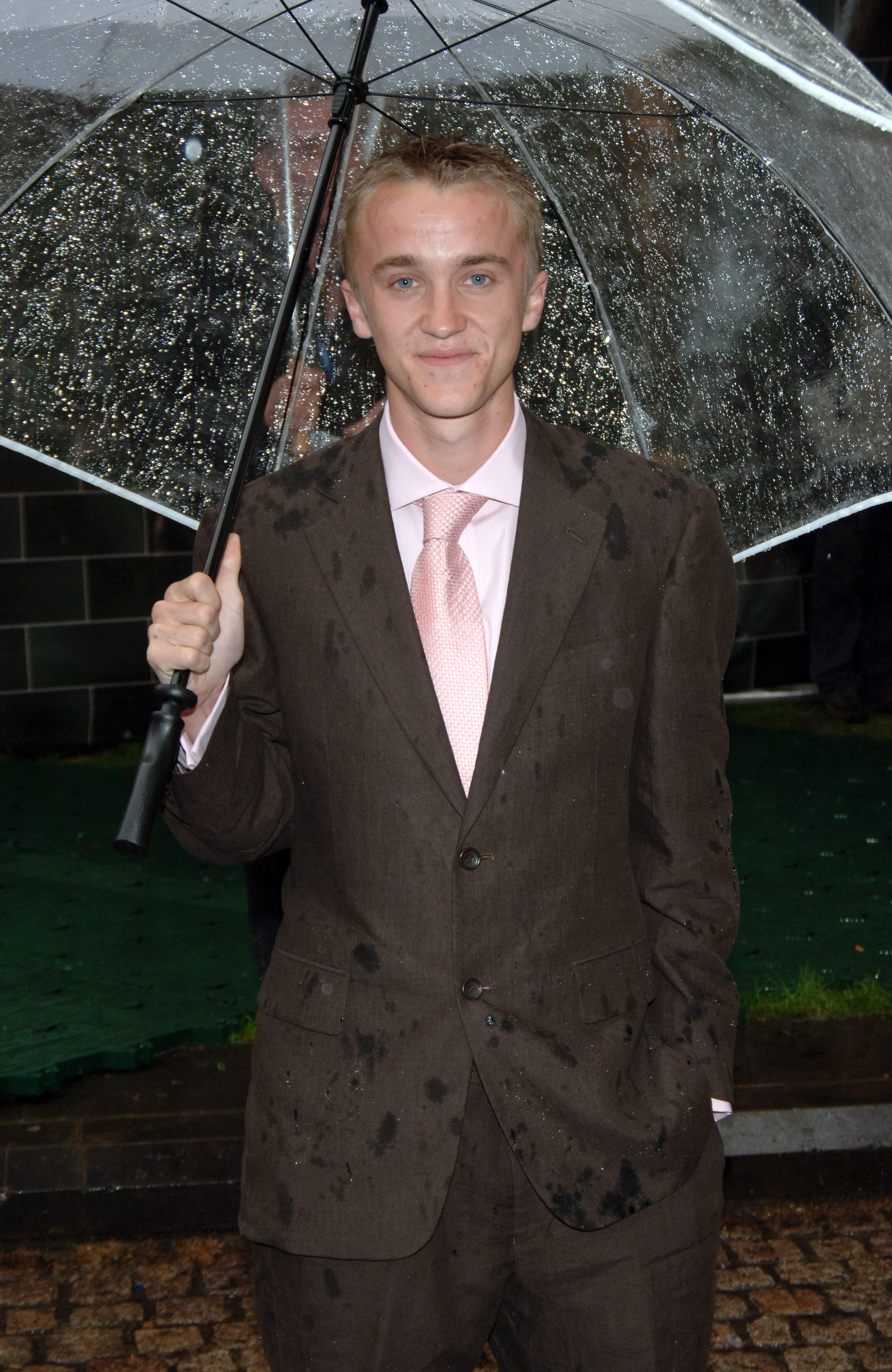 Tom Felton stands under a clear umbrella, rain-speckled and smiling as if the storm were part of the event's charm. His suit bears the marks of London's drizzle, yet he carries himself with easy confidence and good humor. It's a quintessentially British premiere moment and the magic goes on.